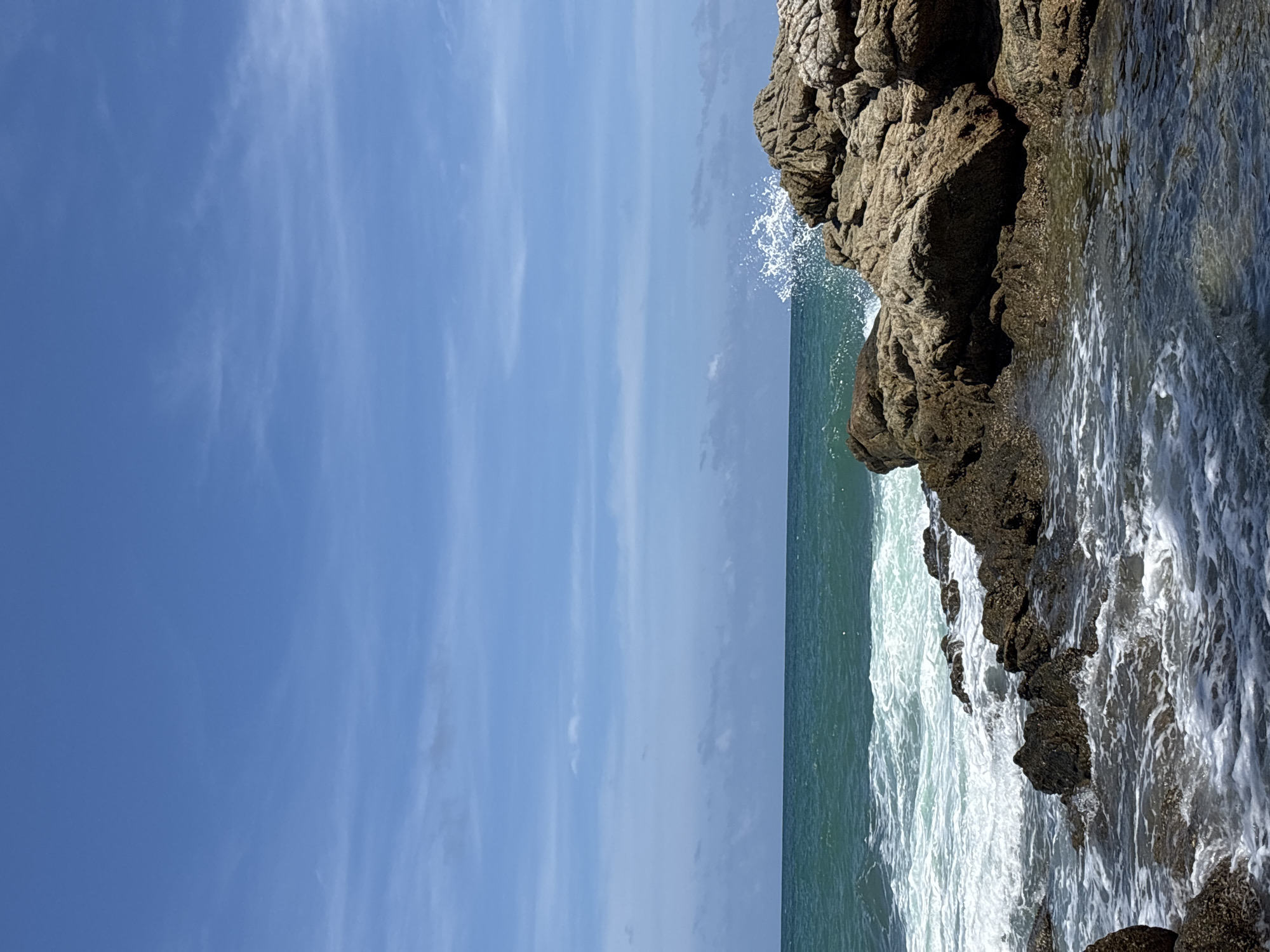 A coastal scene in Choeng Thale, Thailand, features a rocky outcropping on the right, with ocean waves crashing against the rocks. Water sprays into the air at the point of impact. The ocean extends to the horizon, where it meets a sky with light clouds.