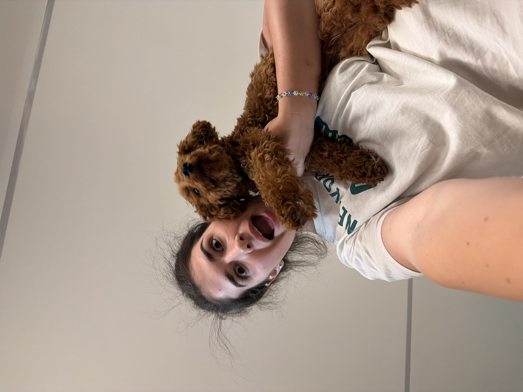 In this upside-down selfie, a young woman with dark hair is holding a small, fluffy brown dog over her face. The dog is positioned in front of her mouth and nose, with its paws resting on her chest. The woman's eyes are wide open, and her mouth is agape in a playful or surprised expression. She is wearing a light-colored t-shirt with dark green text that is partially obscured. On her left wrist, she wears a beaded bracelet with blue, white, and green beads. The background is a plain, light-colored wall, suggesting an indoor setting. The lighting is soft and appears to be from an overhead source. The overall impression is one of lighthearted fun and affection between the woman and her dog. The image was likely taken during the day.
