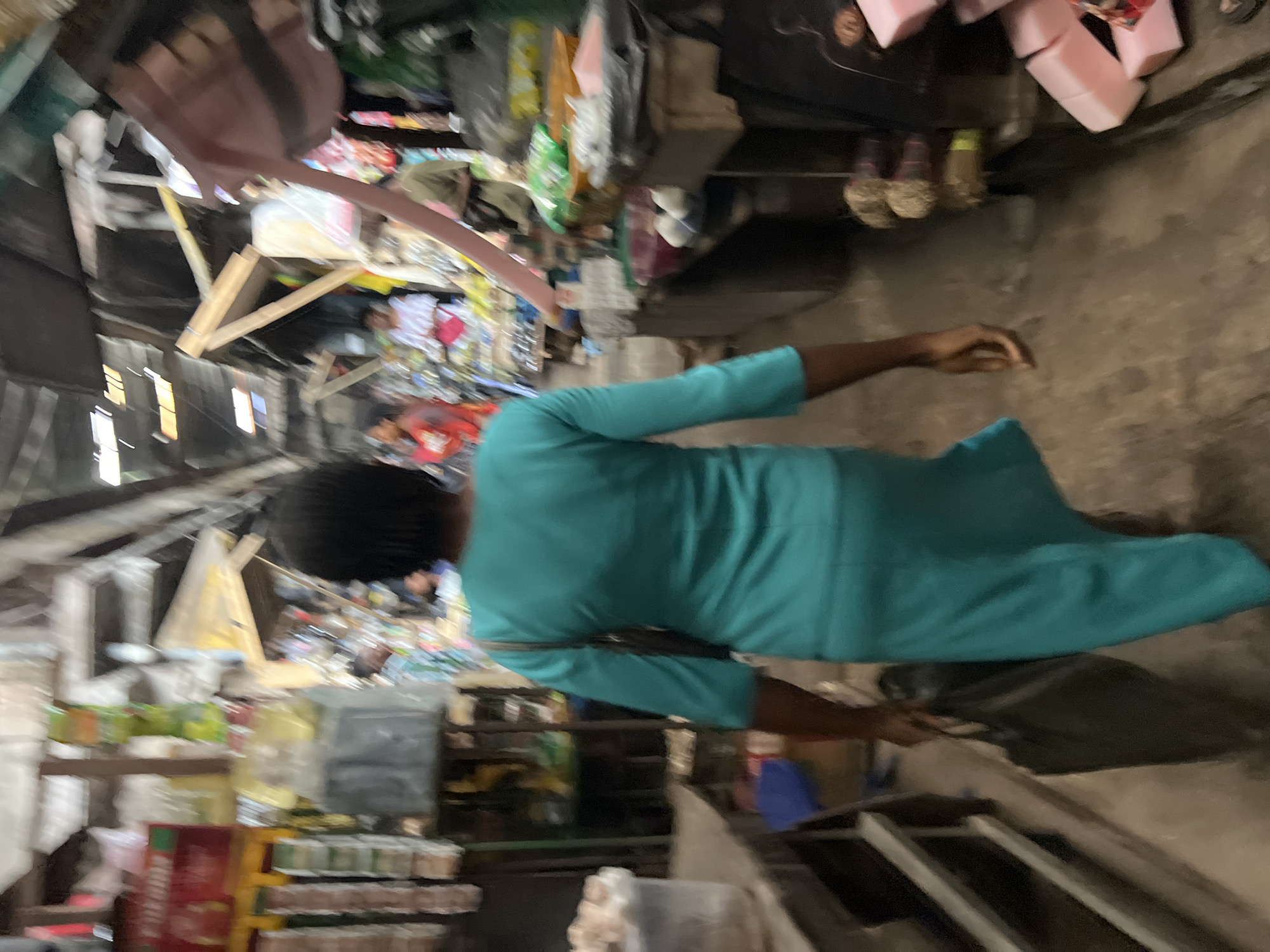 The image depicts a blurred scene of a bustling market in Nkpor, Nigeria. A person in a teal dress is walking away from the camera, carrying a dark bag. The market appears to be an open-air or covered structure with stalls lining a walkway. The stalls are filled with various goods, including packaged items, produce, and other merchandise. Other individuals are visible in the background, some browsing at stalls and others walking through the market. The overall atmosphere suggests a busy commercial environment.