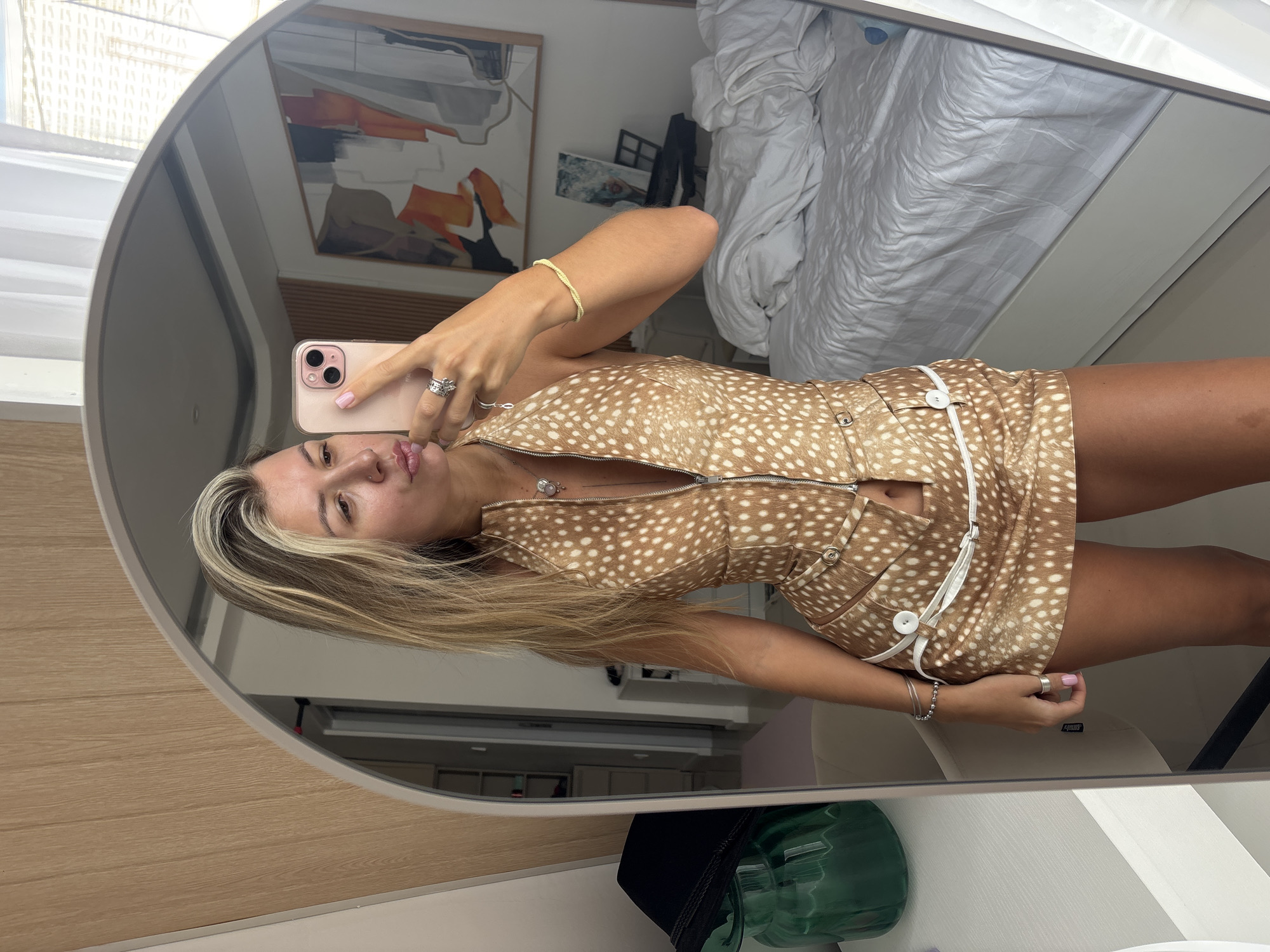 A woman stands in front of a full-length mirror, taking a selfie with her phone. She is wearing a brown and white patterned sleeveless halter top and a matching skirt. The top has a zipper down the front and the skirt has white detailing and buttons. Her hair is blonde and wavy, and she is wearing jewelry on her fingers, neck, and wrist. In the reflection of the mirror, a white bed with rumpled sheets is visible, along with a framed piece of abstract art on the wall. To the left of the woman, a green glass vase with a black hat is on a white dresser. The room appears to be a bedroom.