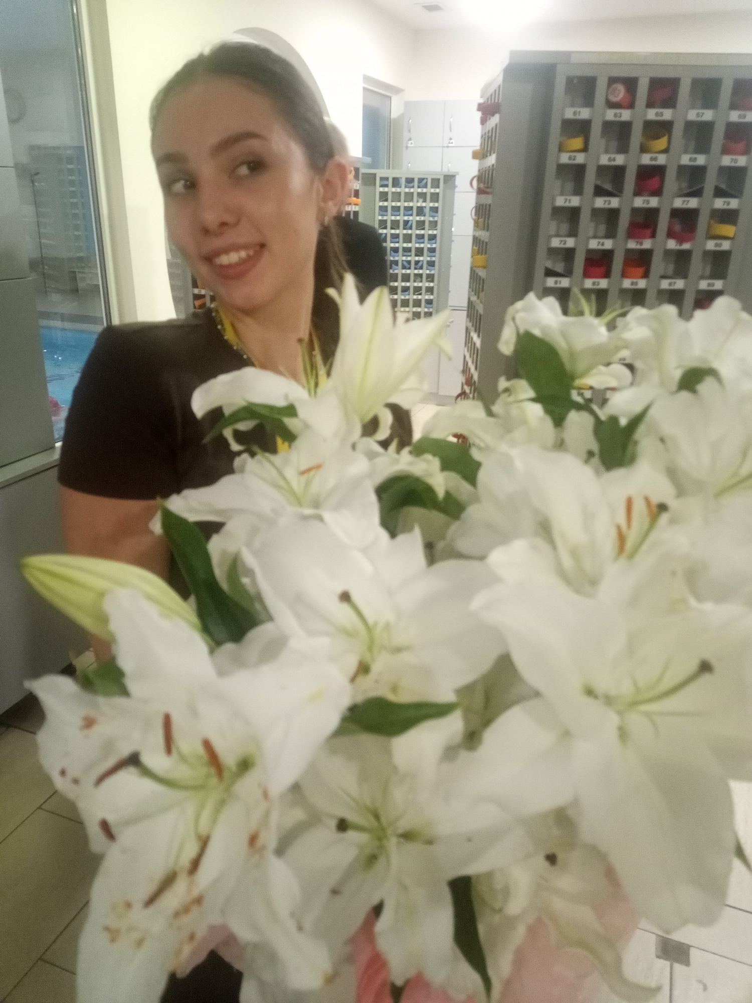 In an indoor setting, a young woman with dark hair tied back smiles at the camera while holding a large bouquet of white lilies. She is wearing a dark t-shirt with a yellow lanyard. To the right of the woman, a grey metal cabinet with numbered compartments is visible. In the background to the left, a swimming pool with blue water can be seen through a large window. The scene appears to be within a public facility, possibly a locker room or a recreational center, given the presence of the lockers and the swimming pool.