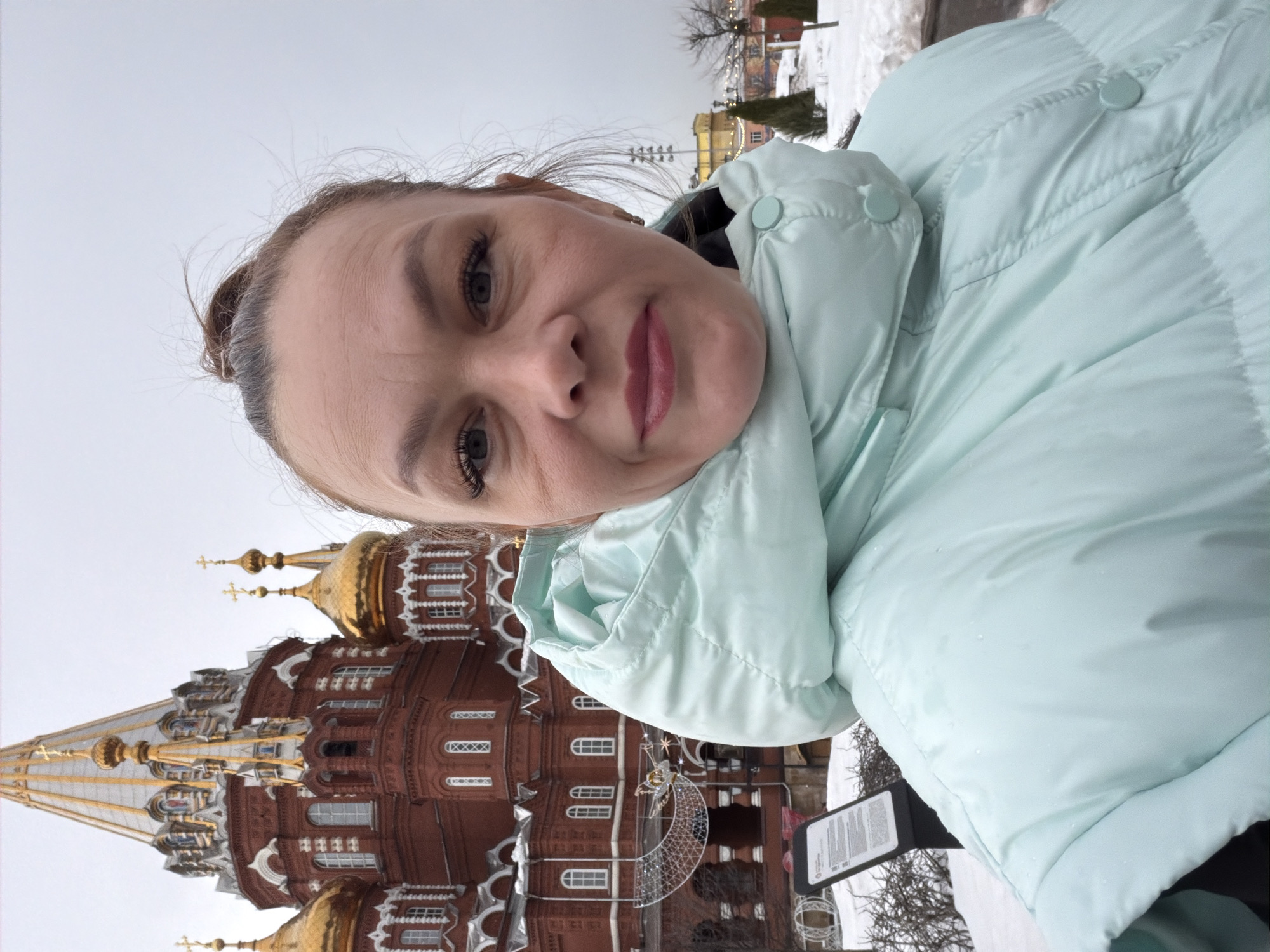 An adult female is depicted in a close-up, selfie-style photograph, positioned centrally and looking directly at the camera. She has her hair pulled back and wears a light blue (mint green) hooded puffy winter jacket, on which small water droplets are visible. Her lips are closed with a slight upward curve.
In the background, the Saint Michael's Cathedral (Mikhailovsky Cathedral) in Izhevsk, Russia, is prominent. This large building is constructed of red brick with white architectural detailing and features multiple golden onion domes, each topped with an Orthodox cross. A large, decorative light fixture, partially shaped like a crescent moon, is visible near the cathedral's base.
The ground is covered in snow. A dark informational sign with white text is visible in the lower-left foreground. The sky is overcast and uniformly grey. In the distant background, bare trees and a yellow building with stadium lights are present.