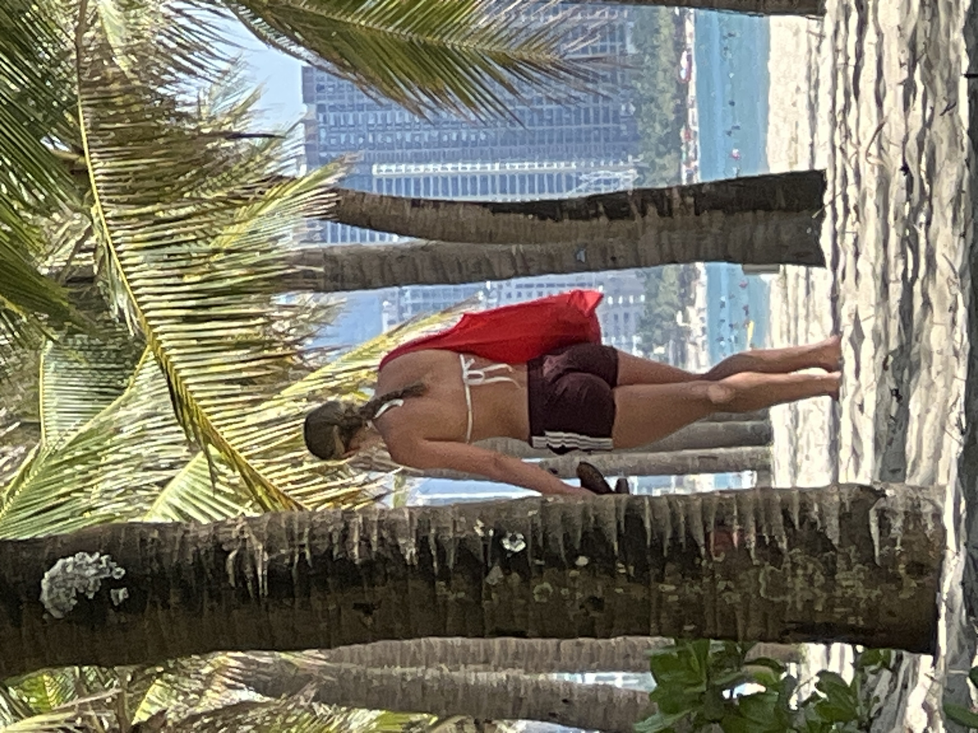A woman is walking on a beach in Da Nang, Vietnam, near the city skyline. She is wearing a white bikini top and maroon shorts with white stripes. She is carrying a red bag over her right shoulder. The beach is lined with palm trees, and the ocean is visible in the background with several boats and swimmers. The city skyline with tall buildings is also visible in the distance. The sand on the beach is light brown.