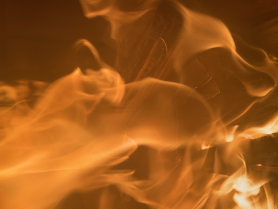 A close-up photograph depicts active flames, located in Sedlartsi, Bulgaria. Dynamic, bright orange and yellow fire occupies the majority of the frame, exhibiting turbulent, upward-moving patterns. Submerged within the flames, dark, irregular fragments consistent with burning wood are faintly discernible, primarily in the central and right portions. The background is dark and featureless. No persons or other identifiable objects are present.