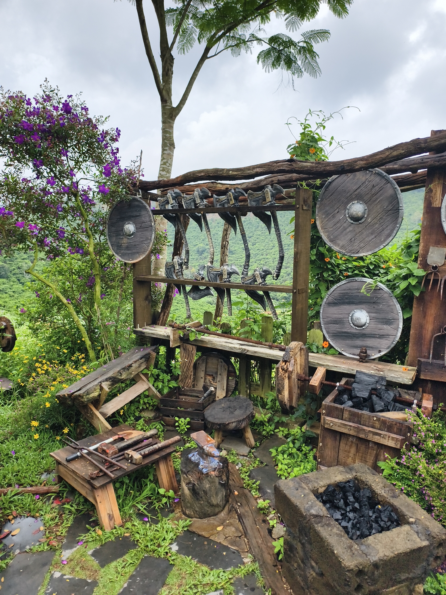The scene depicts a rustic outdoor display, possibly a blacksmith's or craftsperson's workspace, set in Thôn 1, Vietnam. Several wooden structures form the backdrop, featuring shields and axes displayed on shelves. A collection of tools, including hammers, tongs, and metal rods, rest on a wooden workbench. A stump serves as an anvil, showing signs of use. Coal is stored in wooden boxes and a stone-lined fire pit. The area is surrounded by lush vegetation, including purple-flowering shrubs and other plants, suggesting a natural, possibly rural, environment. The background reveals a hillside and overcast sky. No people are visible. The overall impression is that of a staged or maintained historical exhibit or an artisan's workshop integrated into the natural landscape.