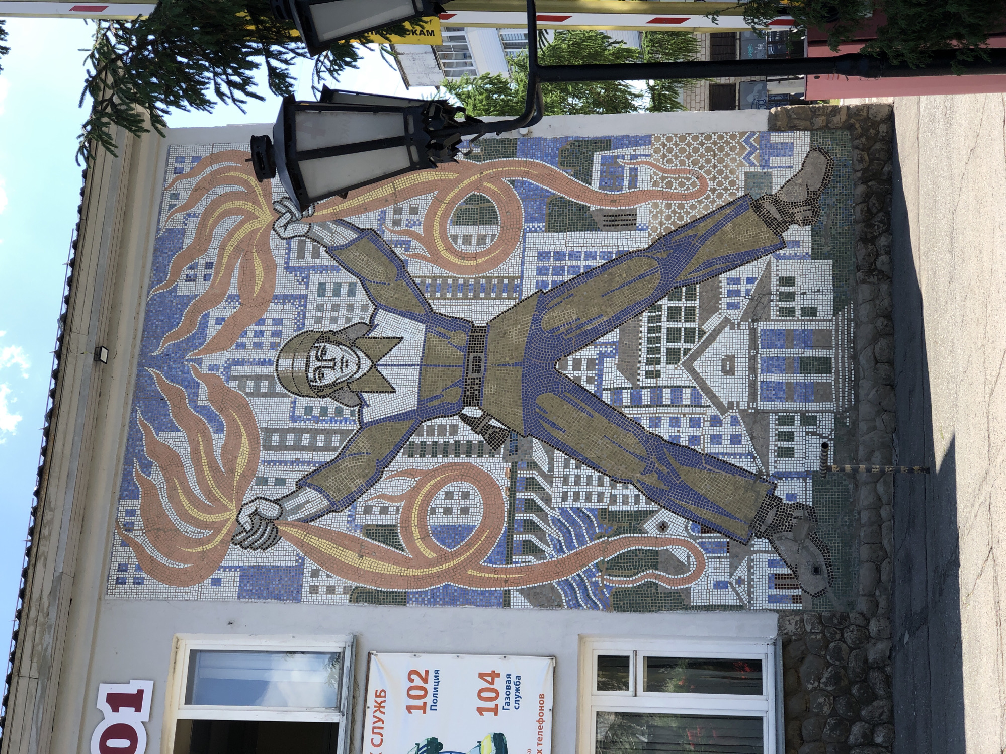 The image shows a mosaic mural on the side of a building in Chaikovsky, Russia. The mural depicts a stylized human figure in a protective suit with arms and legs outstretched. In each hand, the figure holds a representation of flames. A winding design surrounds the figure, and the background shows a city landscape. The building's façade also features windows and signage with the numbers "102" and "104," along with Russian text. A street and lamppost are visible in the foreground.