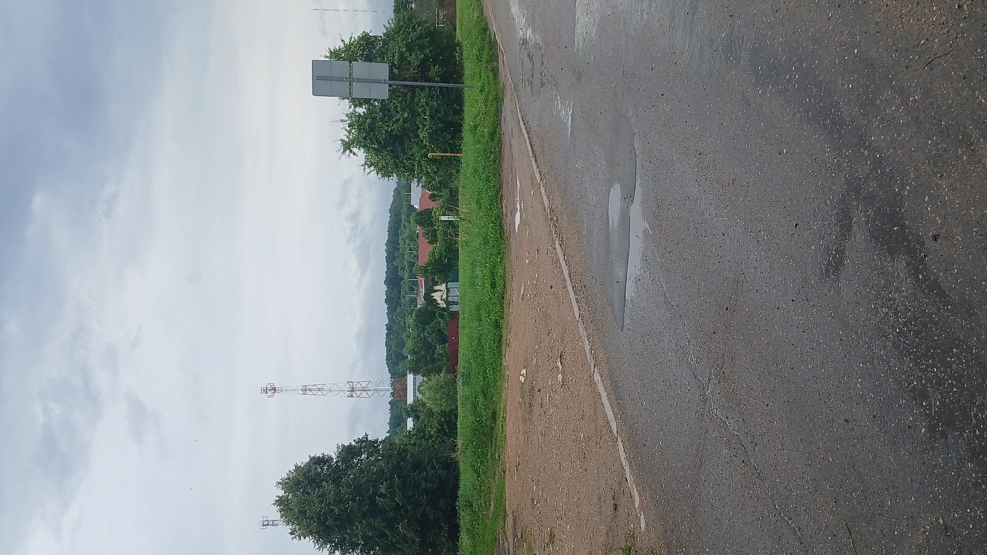 The image depicts a rural or suburban roadside scene under an overcast sky. In the foreground, a wet asphalt road with visible cracks and puddles stretches from the bottom edge towards the right. A dirt or gravel shoulder runs along the left side of the asphalt, separated by a white line. Beyond the shoulder, a grassy embankment leads to a line of trees and buildings in the middle ground. Two tall telecommunication towers, one red and white lattice, and another with antennas, are visible in the distance. A traffic sign with two grey panels stands near a large, leafy green tree on the right side of the middle ground. The background consists of rolling hills covered in lush green trees, with glimpses of red-roofed houses nestled amongst them. The overall atmosphere is calm and depicts a typical day in a non-urban environment, possibly in the vicinity of Ivakino, Russia, given the context. No people or animals are visible in the frame.