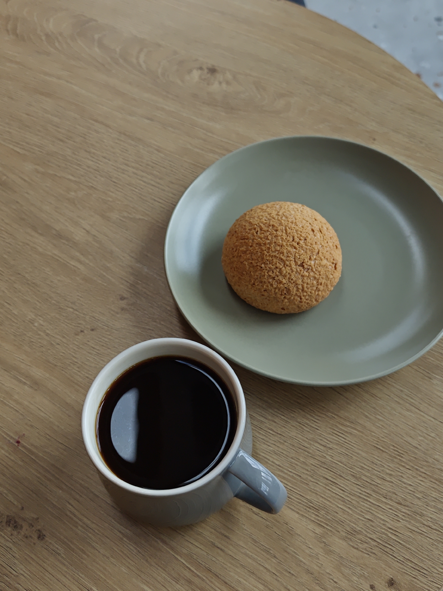 A high-angle shot features a light-brown, wood-grain table surface. A light-grey mug filled with dark coffee is positioned on the lower left. A light-green plate, holding a round, textured, light-brown pastry, is situated on the upper right. The location is unknown and can not be determined from the image. There are no people, actions, or interactions visible in the image.