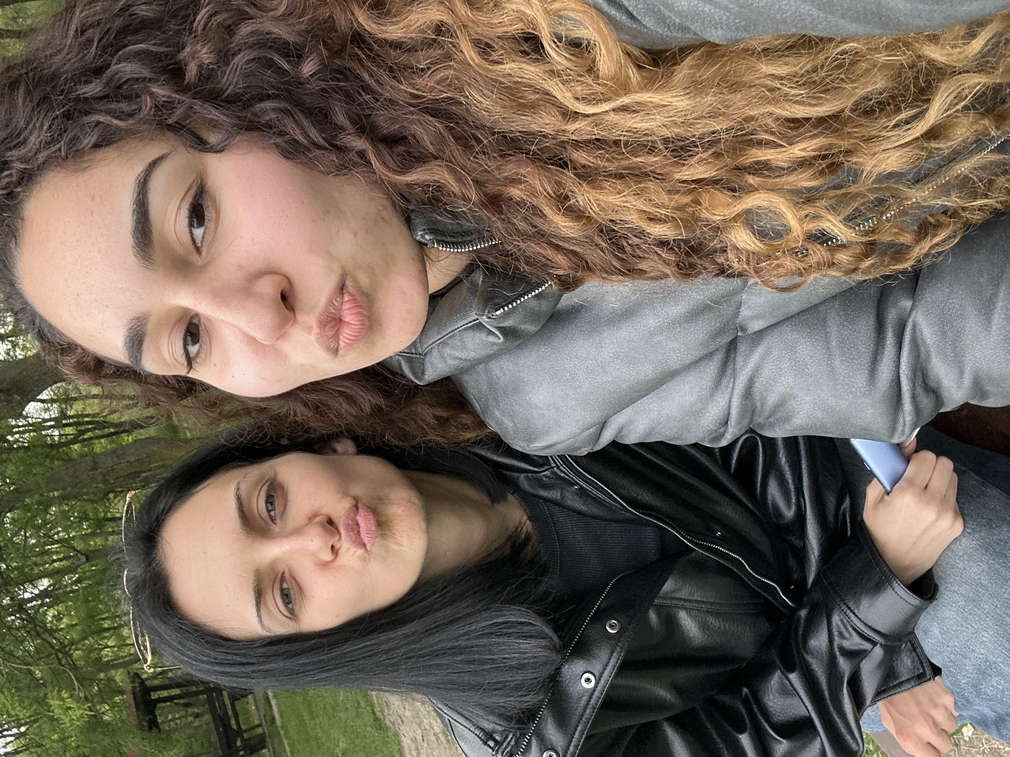 Two women are posing outdoors in what appears to be a park. The woman at the top has curly, brown and blonde ombre hair and is wearing a gray jacket. The woman at the bottom has long, straight black hair and is wearing a black leather jacket. Both women are making a kissing face. The background consists of green foliage and trees. The location is not discernible as being within Burgas, Bulgaria from the visual information.