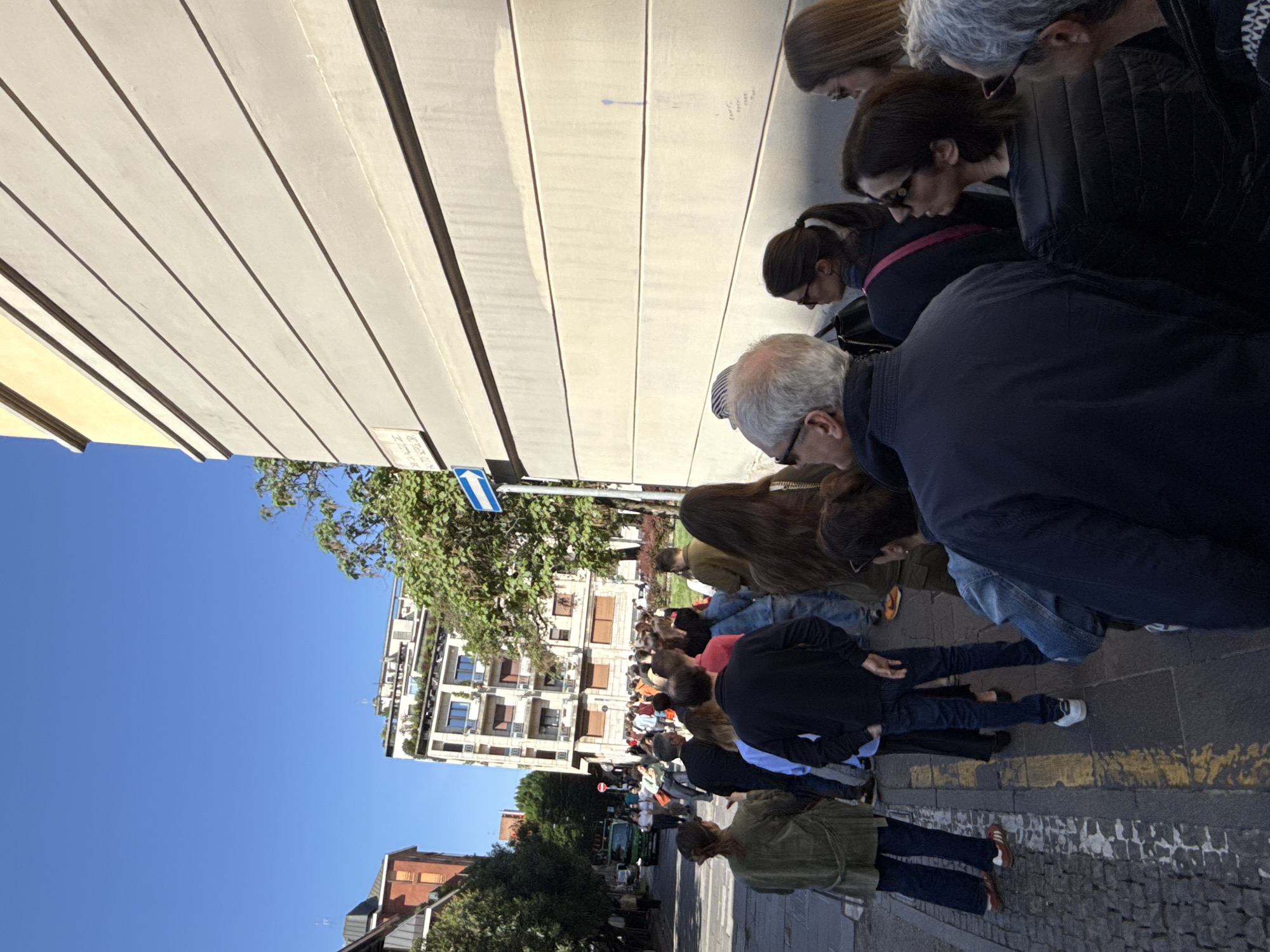 This image, rotated vertically, captures an urban scene in what is specified as Milan, Italy, on a bright, sunny day. A large group of people, primarily adults, are gathered along a long, light-colored segmented wall, bending forward and looking intently at something on its surface. Their backs are mostly towards the viewer, suggesting a common focus on the wall or the ground immediately in front of it. The individuals are dressed casually in jackets, shirts, and jeans.
In the background, under a clear blue sky, are multi-story buildings with balconies and some greenery, typical of a European city. Some older brick buildings and lush green trees are also visible. The ground is paved with dark gray material, possibly cobblestones, featuring yellow painted lines. A blue and white directional street sign with an arrow is partially visible on the left side of the frame (when re-oriented). Further down the street, more people are seen, indicating a queue or a larger crowd.
The activity suggests a public event, perhaps an art installation, a historical marker, or an exhibition that requires close examination. On the light-colored wall, some very faint, unreadable handwritten text or small drawings are barely discernible, along with a faint blue arrow pointing to the right. The bright sunlight and clear shadows indicate it is likely midday or early afternoon. The people appear focused and curious, engaged in observing whatever is laid out or inscribed along the wall.