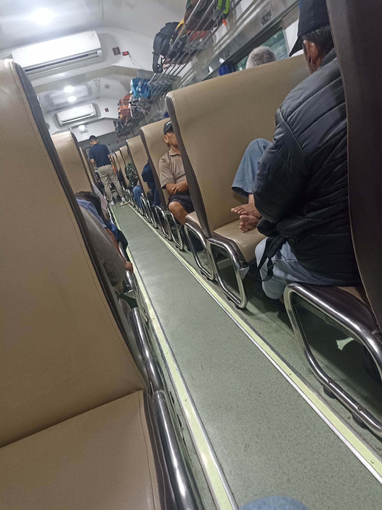 The image depicts the interior of a train carriage, likely in Sumberjati, Indonesia, based on the provided context. The scene is viewed from a low angle, looking down the aisle of the carriage. Several passengers are visible, seated in rows of beige-upholstered seats with chrome frames. In the foreground, a person wearing a black jacket and a dark baseball cap is seated, with their feet exposed and resting on the seat in front. Further down the aisle, another person in a blue shirt is standing and looking forward. A man in a light brown shirt and dark shorts is seated further back, with his arms crossed. Luggage is stowed on overhead racks. Two air conditioning units are visible on the ceiling. The overall environment suggests a mode of public transportation during a journey.