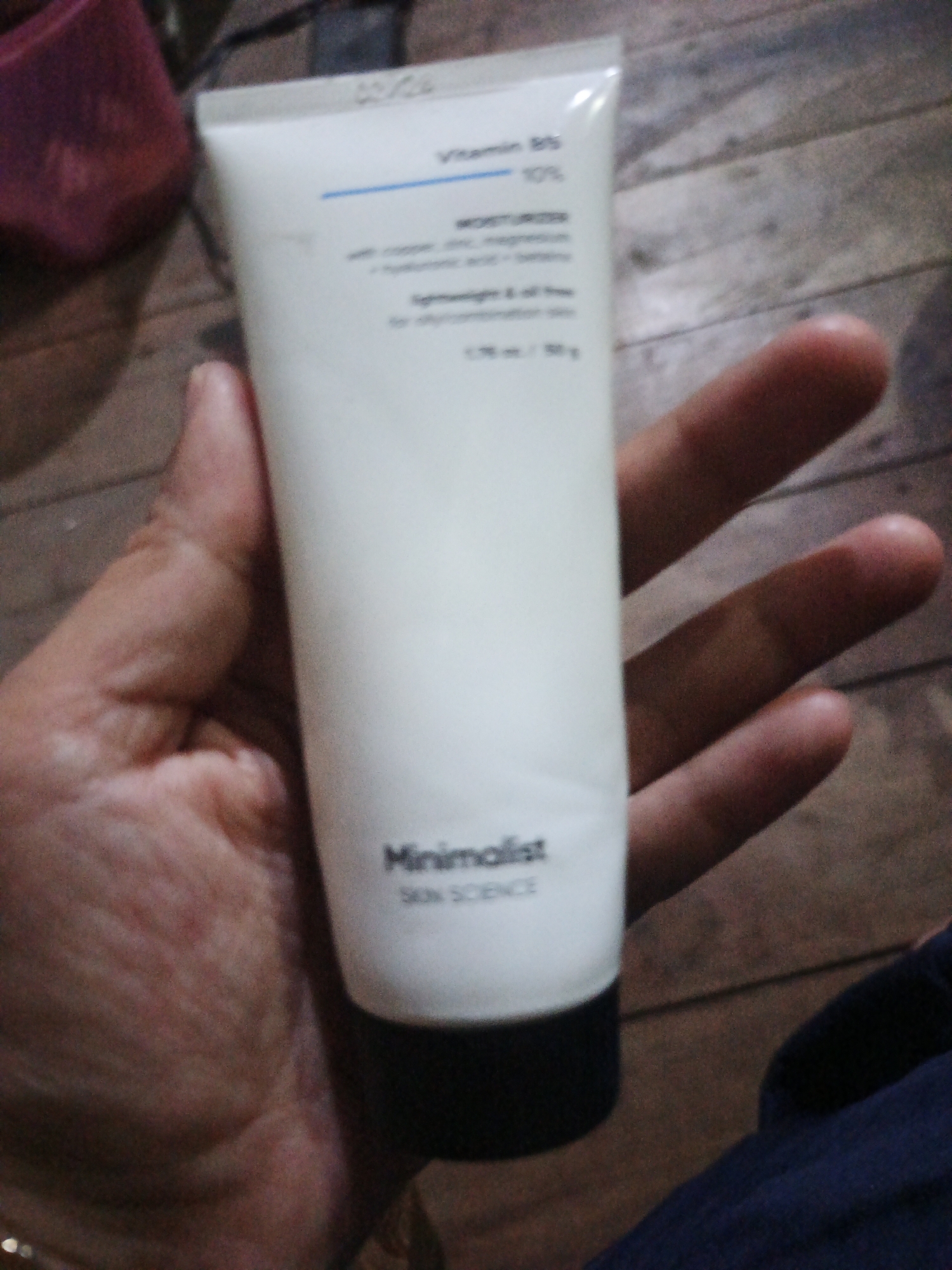 The image shows a hand holding a white tube of "Minimalist Skin Science" moisturizer. The tube has text indicating "Vitamin B5 10%" and describes the product as "Lightweight & oil-free for all skin types." A person's hand is holding the tube, with their fingers partially obscuring the product. The background appears to be a wooden surface. No specific location in Sinar Bulan, Indonesia, is discernible from the image.