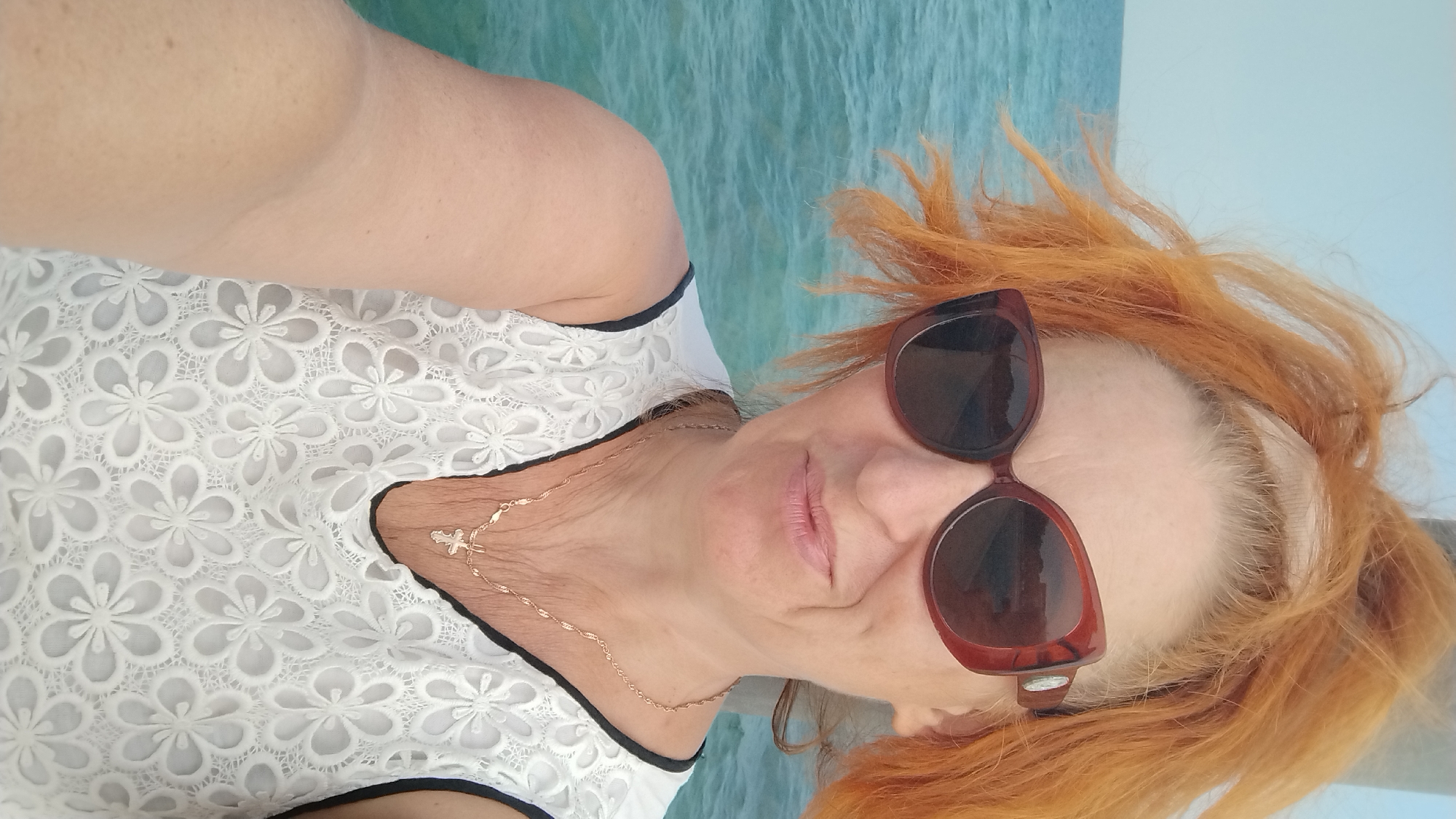 A woman with vibrant orange hair, wearing large brown sunglasses, a white lace top, and a gold necklace with a cross pendant, smiles at the camera. She is positioned in front of a body of turquoise water, suggesting an outdoor setting, possibly near the sea. The background consists of the clear blue sky and the rippling surface of the water. The image has a selfie-like composition, with the woman's left arm visible in the foreground. The overall impression is of a candid moment captured in a sunny, coastal environment. The provided location, Perea, Greece, aligns with this interpretation, as Perea is a coastal town in Thessaloniki.