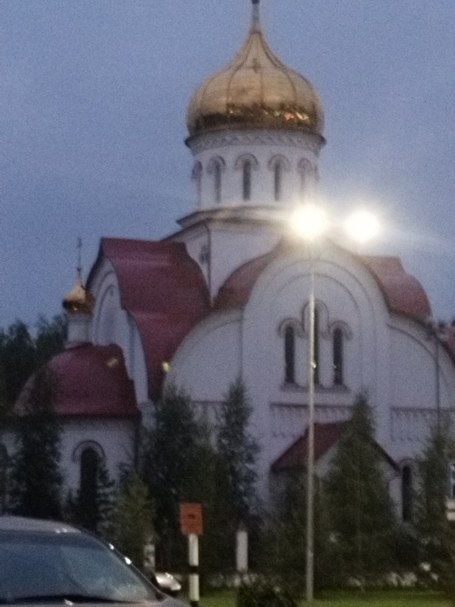 The image shows a church with a golden dome and red roofs. The building is white and appears to be made of stone or concrete. There are several arched windows on the building. In front of the church, there is a street lamp with two lights. The sky is overcast and the lighting suggests it is dusk or dawn. There are trees and bushes around the church. The foreground shows the hood of a car, indicating it is parked or driving past the church. The building appears to be the Church of the Assumption of the Blessed Virgin Mary in Surgut, Russia, as it is a prominent landmark with a golden dome and red roofs, consistent with the architecture shown.