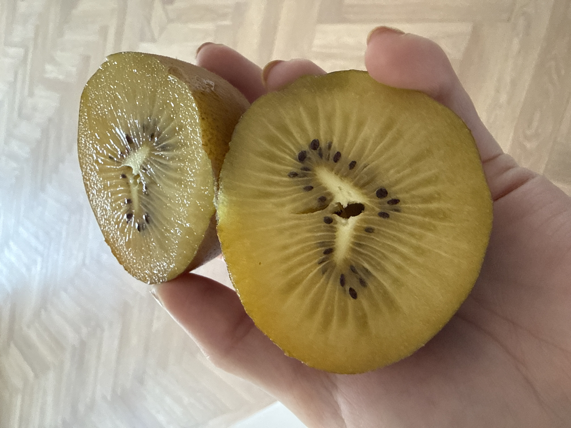 A hand holds two halves of a cut golden kiwi fruit. The fruit's flesh is yellow with black seeds radiating from the center. The background is a blurred, light-colored surface with a faint pattern. The city and country are not discernible.