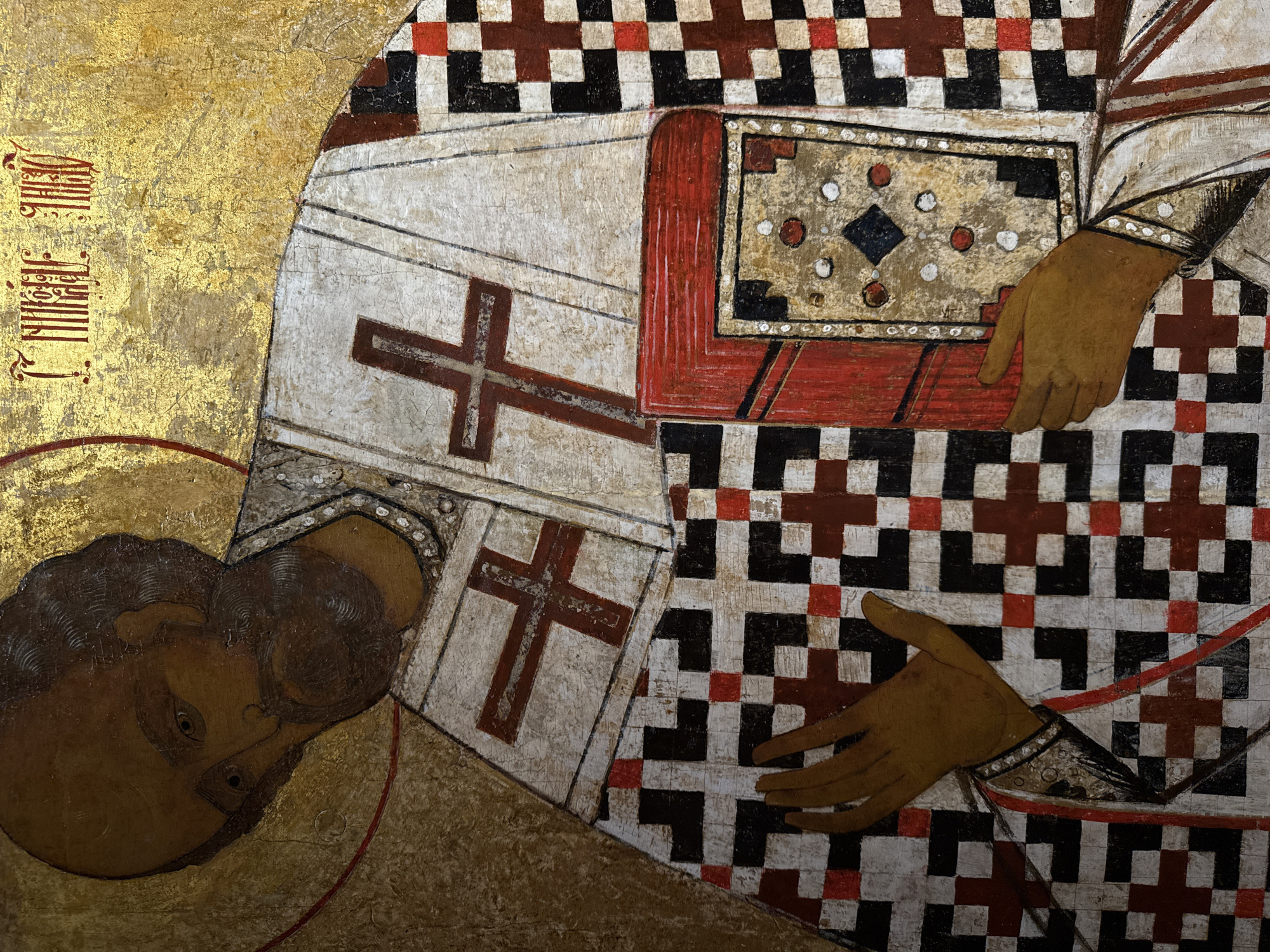 This is a detail of an Eastern Orthodox icon depicting the beheading of St. John the Baptist, specifically a fragment of the icon found in the Church of St. John the Baptist in Nizhny Novgorod, Russia. The lower left corner shows the head of St. John, laying at an angle. A portion of a red halo is visible around his head. To the right, a larger figure is shown in partial view, possibly representing the executioner. This figure is adorned in a white garment with red crosses and checkered patterns of black, white, and red squares. The executioner holds the head of the saint.