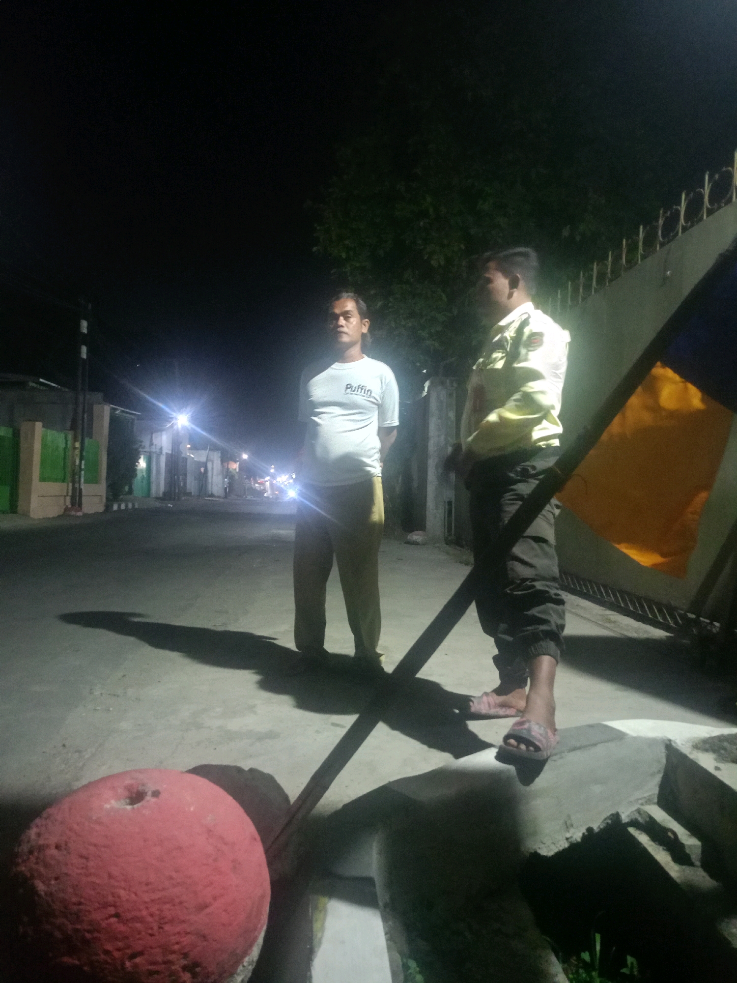 Two men stand on a paved street at night in Cirebon, Indonesia. The man on the left wears a white t-shirt with the word "Puffin" on it and light brown pants. He casts a long shadow on the ground. The man on the right wears a light yellow collared shirt and dark pants. He is partially obscured by a dark pole and a bright orange flag. In the foreground, a large, textured red spherical object rests on the ground. The background shows buildings, streetlights, and trees, suggesting a residential or commercial area.