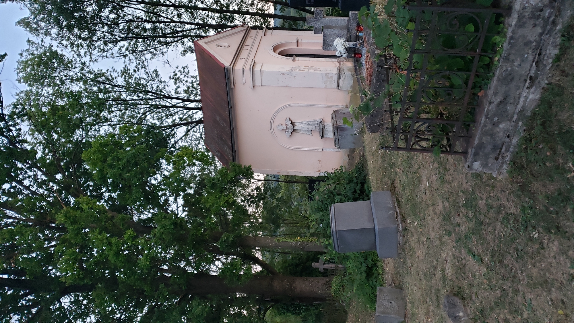 The image depicts a cemetery scene in Prešov, Slovakia. In the foreground, a weathered grey tombstone is visible, with a decorative metal fence partially obscuring a cluster of green foliage behind it. To the left, a tall, dark grey tombstone stands prominently. Further back, a light pink mausoleum with a red roof is the central structure, featuring an arched entrance and a relief sculpture above it. A stone statue of a cross-bearing figure is placed in front of the mausoleum. The surrounding environment consists of dry grass, scattered rocks, and a dense canopy of trees, suggesting an outdoor, possibly overgrown, cemetery setting. The lighting indicates it is either early morning or late evening.