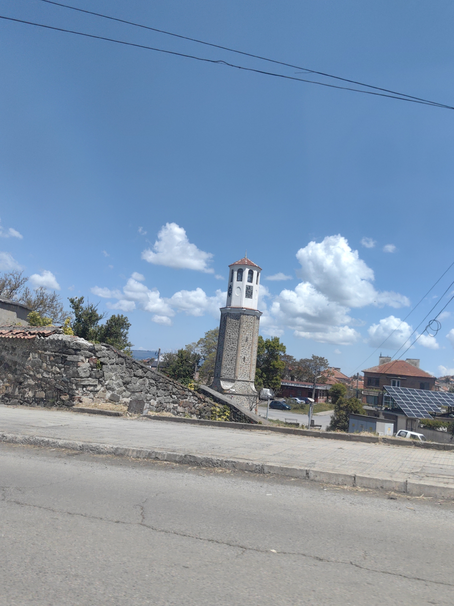 The image depicts the clock tower in the city of Karnobat, Bulgaria. The clock tower is made of stone and has a red tile roof. It is located on a small raised area next to a stone wall, with a sidewalk and road in the foreground. In the background, there are several buildings, trees, and a bright blue sky with scattered clouds. The solar panels are present on the rooftop of the building in the background. Power lines are visible overhead. There are no people visible in the image.