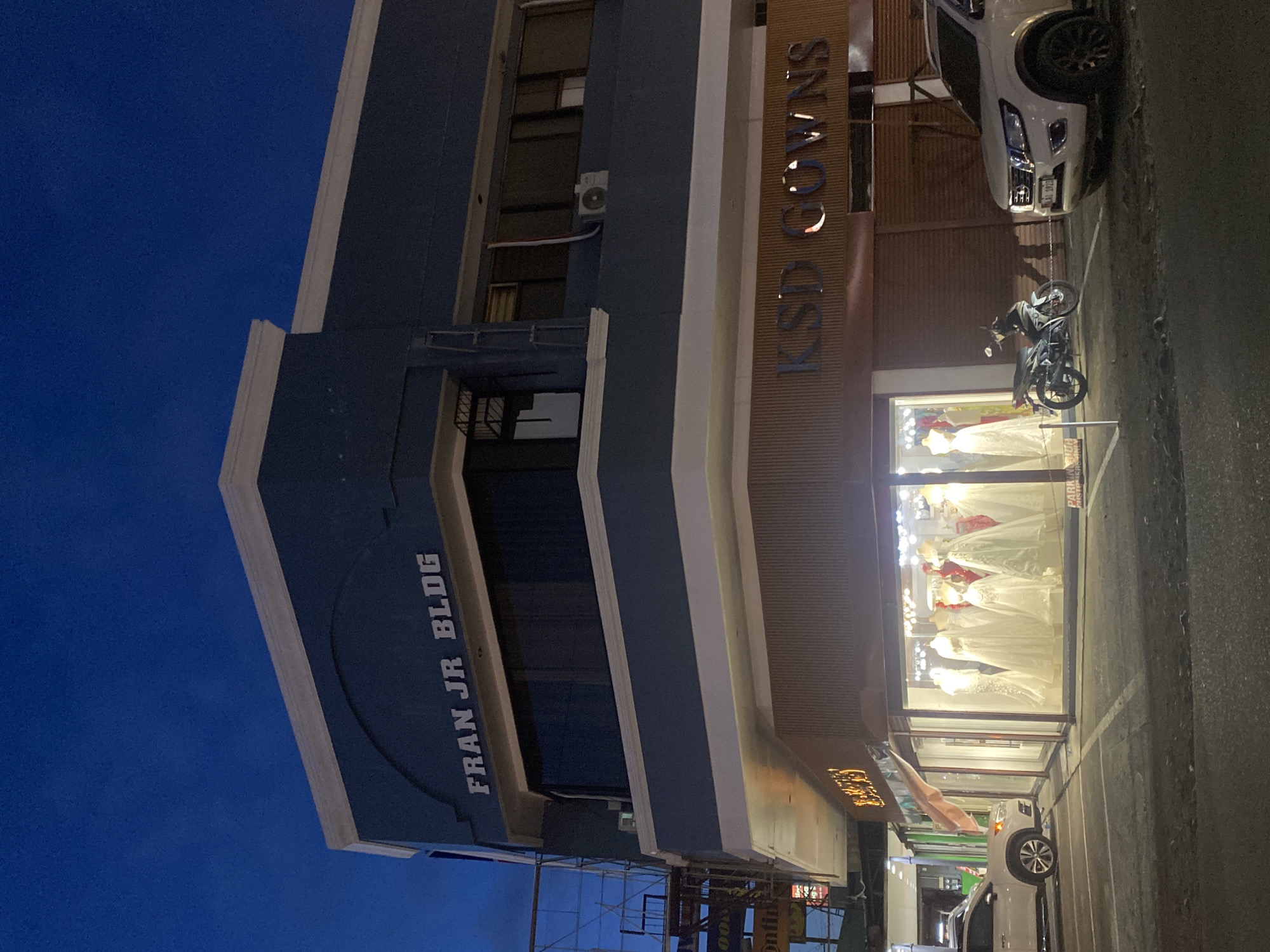 This late evening or nighttime image depicts a multi-story commercial building on a street in Davao City, Philippines. The sky above is a deep blue, indicative of twilight or night, with no stars visible. The building's exterior is painted a dark blue-grey with white cornices and trim. Prominently displayed on the upper facade is the white text "FRAN JR BLDG".
The ground floor of the building houses "KSD GOWNS," identified by illuminated bronze-colored lettering above its display windows. The storefront on the left side features several white and off-white wedding or formal gowns displayed on mannequins, brightly lit from within. To the right of the gown shop's visible entrance, a section of the ground floor is covered by a closed, brown roll-down security shutter.
Three vehicles are present: a white SUV (Nissan Navara or similar) is parked on the right side, facing left, partially in a designated parking space. Its Philippine license plate, "LAI 1923," is visible. A silver and black motorcycle is centrally parked directly in front of the KSD GOWNS storefront, also within a marked parking space. A third white vehicle, possibly an SUV or crossover, is partially visible on the far left, also parked. The street surface is asphalt with white painted parking space lines. No individuals are visible in the scene. An air conditioning unit is mounted on the second floor of the building, and a metal structure, possibly part of a sign or scaffolding, is partially visible on the far left side of the building.