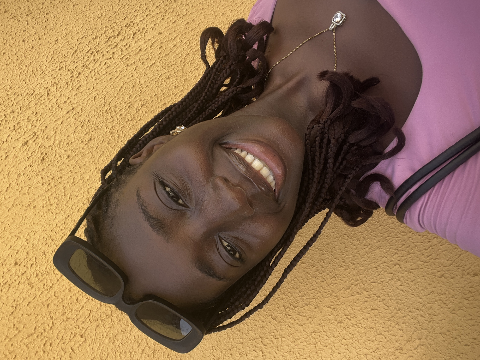 A medium shot captures a dark-skinned woman with dark braided hair, wearing black-rimmed sunglasses, a light purple top, and a gold necklace. She is smiling, showing her teeth. The background is a textured yellow wall. The location is likely in Abuja, Nigeria.