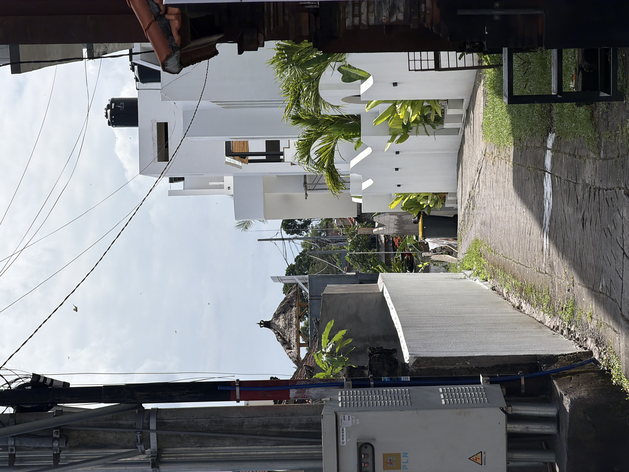 The scene depicts a narrow street in Mengwi, Indonesia. A white, modern-style building with a black water tank on top is visible on one side. Palm trees adorn the facade of the building. The street is paved with concrete and has grass growing along the edges. Power lines and an electrical box with the logo of Perusahaan Listrik Negara (PLN), the Indonesian state electricity company, are in the foreground. A traditional thatched roof structure is partially visible behind the electrical box. The sky is overcast. No people are visible in the frame.