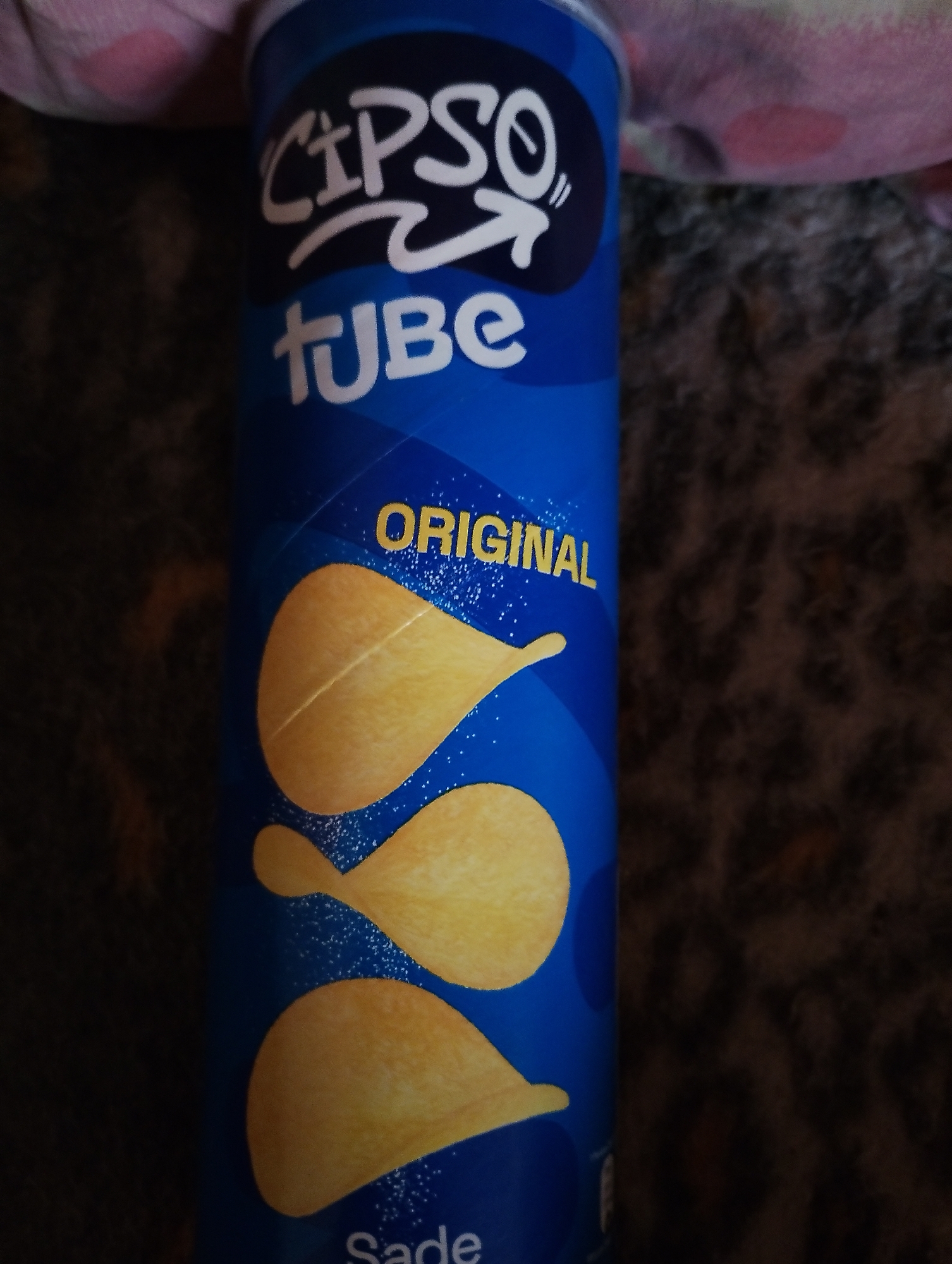 The image shows a blue cylindrical can with the brand name "CIPSO" in white bubbly font and "tube" in white block letters below it. The word "ORIGINAL" is written in yellow block letters across the can. Three yellow potato chips are depicted on the can. The can is placed on a dark, textured surface, possibly a blanket or carpet, and a pink and white polka-dotted fabric is visible in the upper left corner. The scene is indoors, with no discernible urban or environmental context.