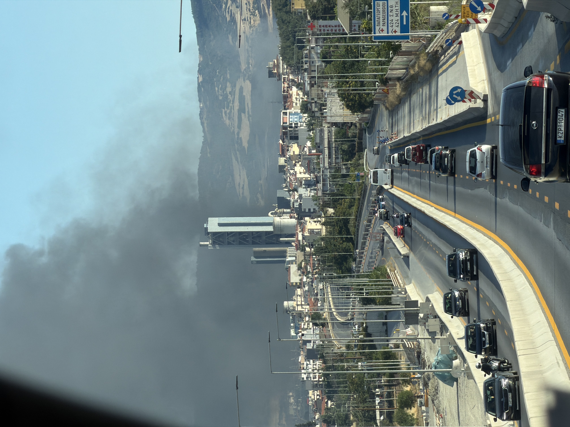 The image depicts a city scene in Polichni Municipal Unit, Greece, dominated by a thick plume of dark smoke rising from behind a cluster of buildings and extending towards the sky. The smoke appears to originate from a location in the background, possibly a fire or industrial emission, obscuring the upper portions of distant hills. In the foreground, a multi-lane highway is visible with several vehicles, including cars and a bus, driving on it. The highway is flanked by concrete barriers and streetlights. Buildings of various sizes and architectural styles are present in the midground, with some appearing to be residential and others potentially industrial, given the presence of tall structures resembling chimneys or towers. A prominent, tall, slender blue and gray building stands out amongst the other structures. The sky is clear and blue above the smoke plume. Signs are visible along the highway, with one blue sign indicating directions to "PAPAGEORGIOU" and "424 Σ.Ν.Θ.". The overall atmosphere is one of urban activity juxtaposed with a significant environmental event indicated by the smoke.