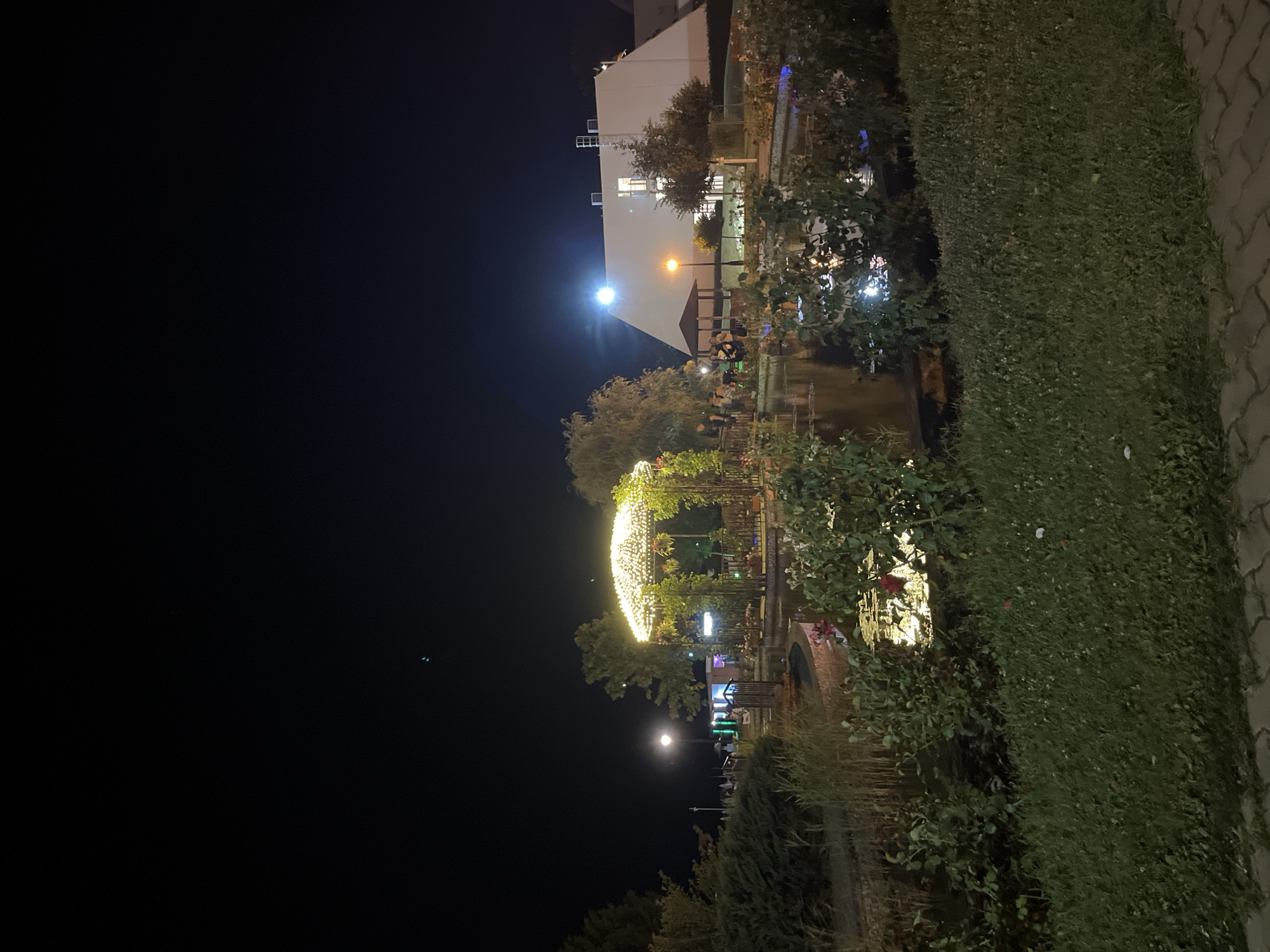 The image depicts a nighttime scene at a park-like area, possibly a waterfront or recreational space, in Obzor, Bulgaria. A brightly lit, decorative gazebo with string lights is the central focus in the mid-ground, situated near a body of water. The water reflects the lights from the gazebo and other surrounding illumination, creating a shimmering effect. On the far side of the water, a large, white, modern building with several windows is visible, illuminated by a bright light source on its exterior. Adjacent to the building, there is a smaller, dark-colored gazebo structure.
Several individuals are present in the scene, some gathered near the water's edge and the structures, suggesting social activity. To the left of the gazebo, there are bushes and trees, and further left, a lamppost provides ambient lighting. The foreground consists of a grassy area and a paved pathway made of interlocking bricks. The overall atmosphere is one of evening leisure and illumination.