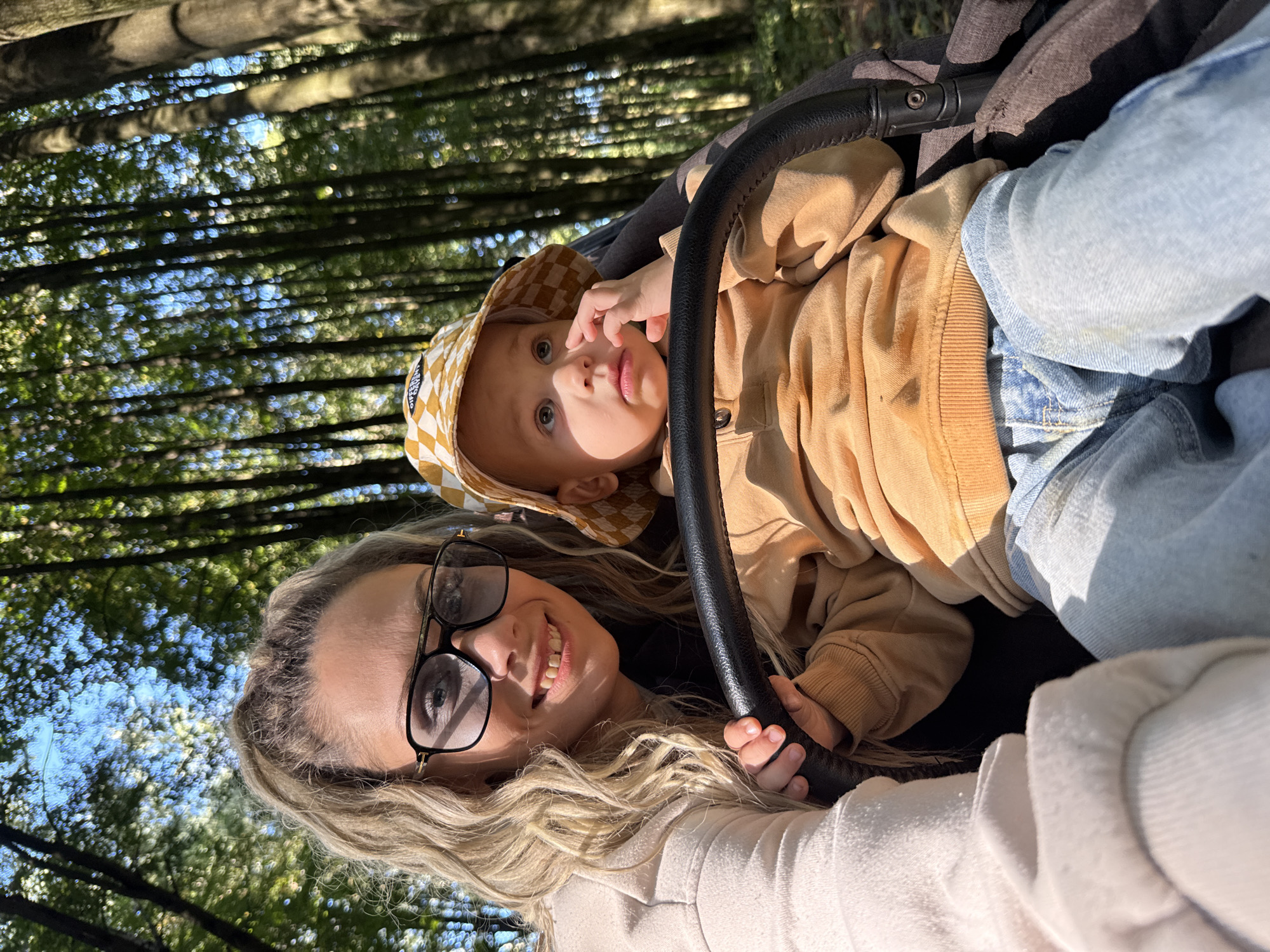 In Rusanovo, Russia, a woman and a baby are in a stroller in a forest. The woman is in the foreground, wearing glasses and a light-colored jacket, with her hand on the stroller's handlebar. The baby, wearing a bucket hat and a tan sweatshirt, is to her right, also in the stroller. Sunlight filters through the trees, illuminating the scene.