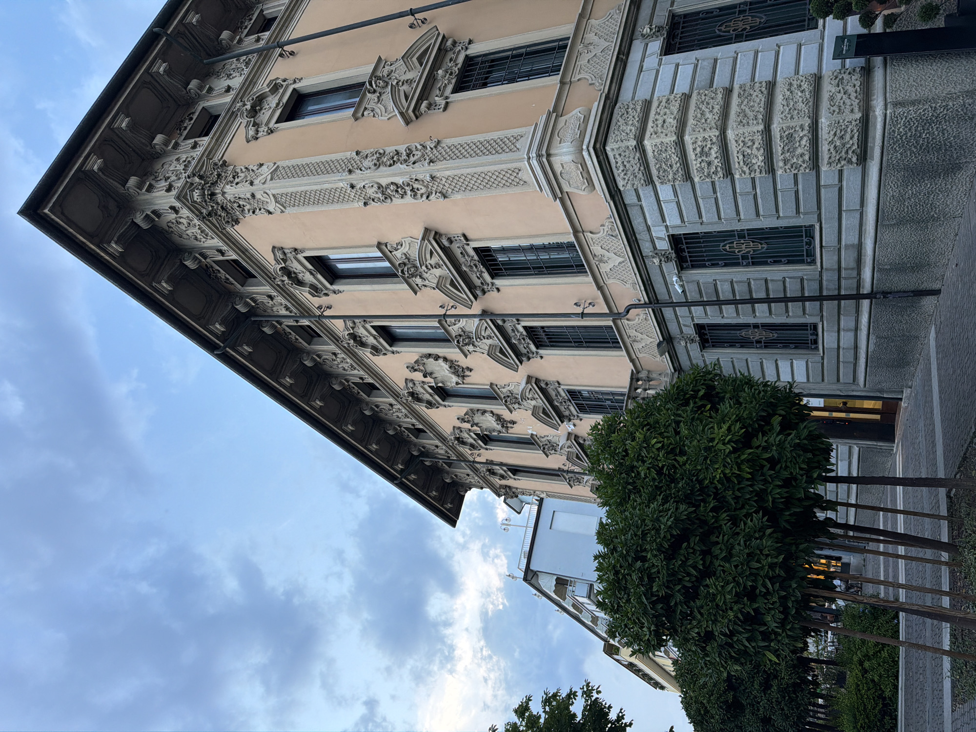 An exterior, low-angle, medium shot captures the corner of a building with ornate, peach-colored upper floors and a grey stone lower floor. The building features intricate architectural details, including decorative moldings, sculpted embellishments around the windows, and a prominent cornice. Multiple windows with dark frames and security grates are visible. In the foreground, several evenly spaced, round, green trees are planted along a paved walkway. Behind the building, another, taller, lighter-colored building is partially visible under a cloudy sky. The scene appears to be an urban environment in Salò, Italy.