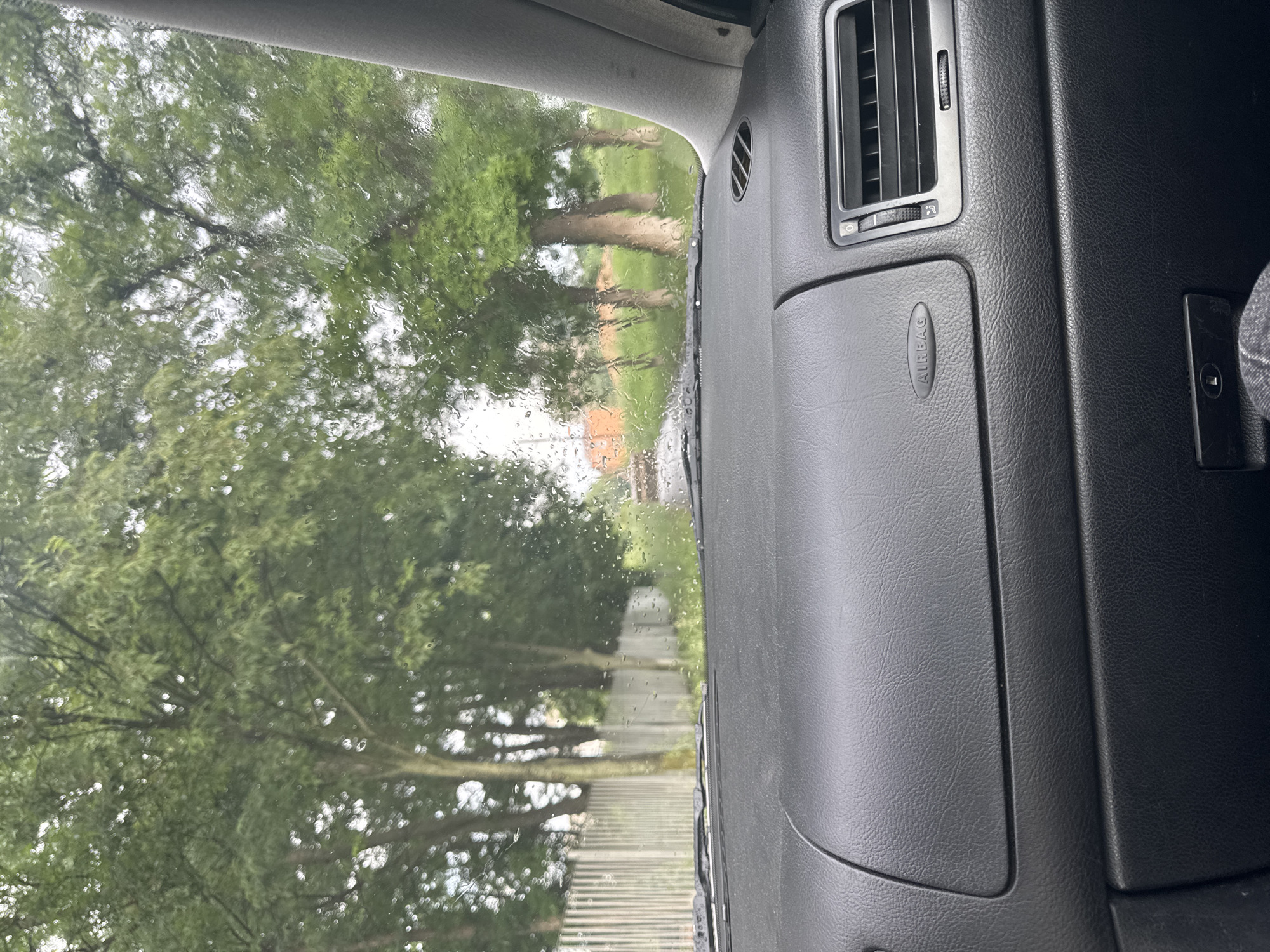 The image captures a view from inside a car, looking out through a rain-streaked windshield on the passenger side. The car's dashboard is prominent on the right, featuring a dark, textured plastic surface. An air conditioning vent with adjustable slats is visible towards the top right, and below it, a cover with the word "AIRBAG" embossed on it. A small, dark rectangular compartment or control panel is also seen further to the right. The upper part of the car's interior, including the roof lining and the passenger door frame, is visible along the top and right edges.
Through the wet windshield, a lush green outdoor scene is discernible, suggesting an overcast day with recent or ongoing rainfall. Numerous water droplets cling to the glass, blurring and distorting the exterior view. The main subjects outside are tall trees with dense green foliage, lining what appears to be a path or a road. The tree trunks are light brown, contrasting with the dark green leaves. In the mid-ground, a paved or unpaved path stretches into the distance, with a faint glimpse of what might be a building or structure (possibly red-brown) further back. The overall impression is of a quiet, possibly rural or park-like setting in Radnevo, Bulgaria, experienced from the shelter of a vehicle during rainy weather.