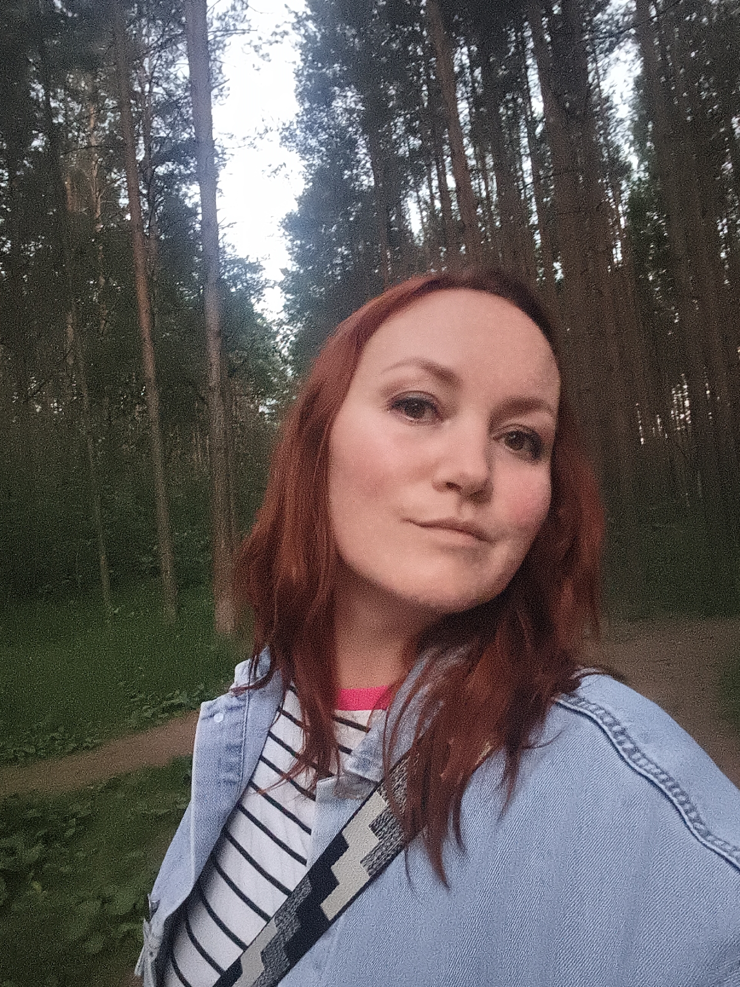 This is a selfie of a woman with reddish-brown hair and fair skin. She is wearing a light blue denim jacket over a striped t-shirt with black and white stripes and a bright pink neckline. A patterned strap, possibly from a bag, is visible across her chest. She has makeup on, including eyeliner and blush. Her expression is neutral, with a slight smile.
The background consists of a forest with tall, slender trees. The lighting suggests it is late afternoon or early evening, as the sky visible through the trees is bright but diffused, indicating sunset or a cloudy day. The ground appears to be a dirt path or trail, with some green grass and foliage on either side. The overall setting is natural and outdoors, likely a park or wooded area. There is no visible text in the image. Given the location context of Chaikovsky, Russia, this scene could be a walk in a local forest or park.