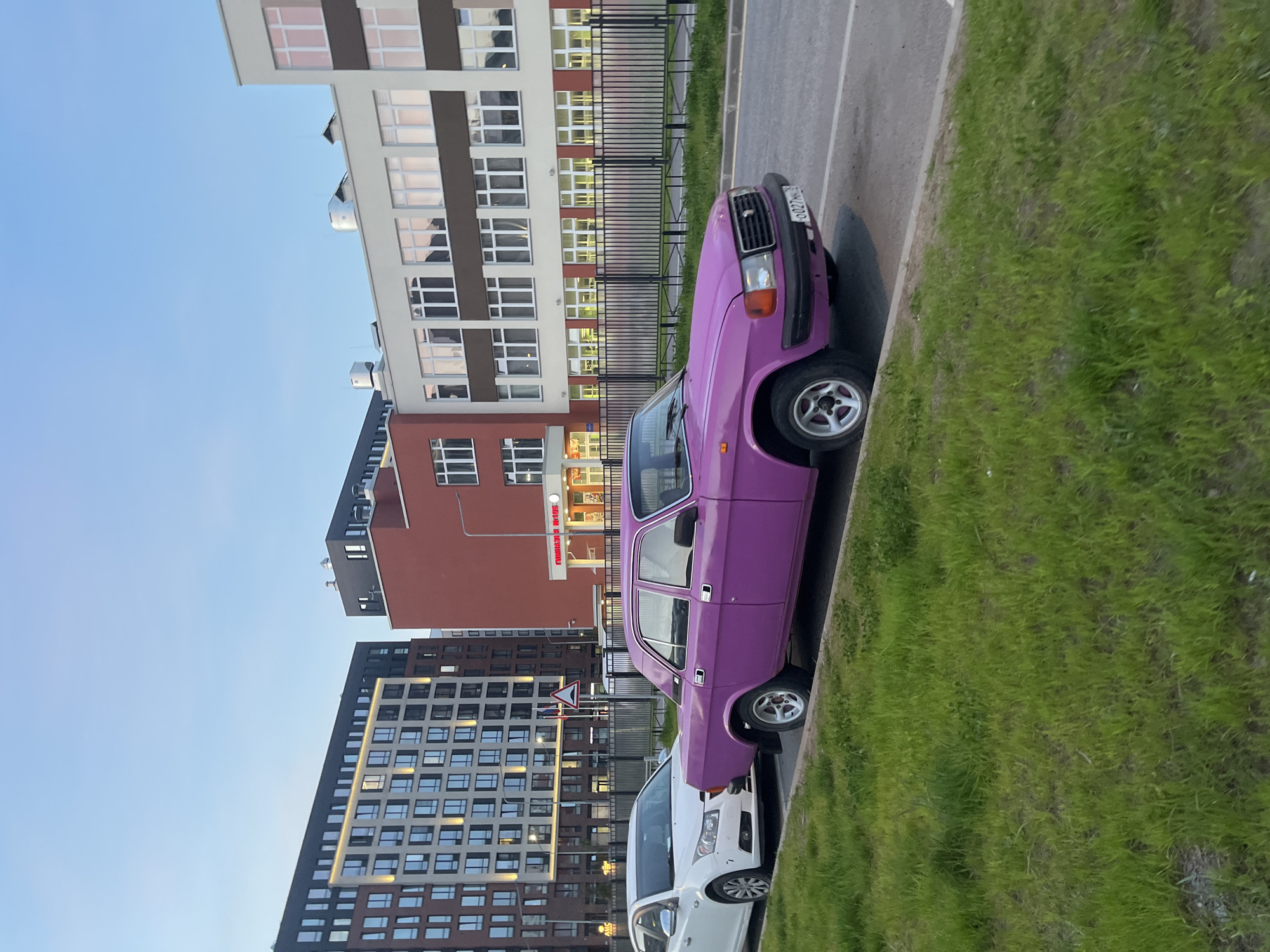 The image depicts a street scene in what appears to be a newly developed area of Saint Petersburg, Russia. A bright purple GAZ-24 Volga car is parked on the right side of the road. In front of it is a white car. The backdrop features several modern multi-story residential buildings of different architectural styles. There is a black metal fence separating the road from the buildings. The sky is blue. The Volga appears to have a license plate with the Russian flag. The grass is green, and the pavement is grey.