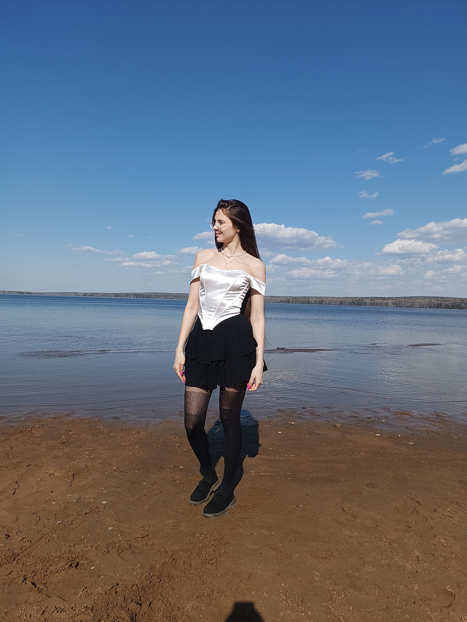 A young woman stands on a beach next to a body of water under a blue sky with scattered clouds. She is wearing a white off-the-shoulder corset top, a short black ruffled skirt, patterned black tights, and black shoes. She has long brown hair and is wearing glasses and a necklace. She is looking to her left. The beach is sandy, and the water stretches out to a tree-lined horizon. The location appears to be near Unknown, Unknown.
