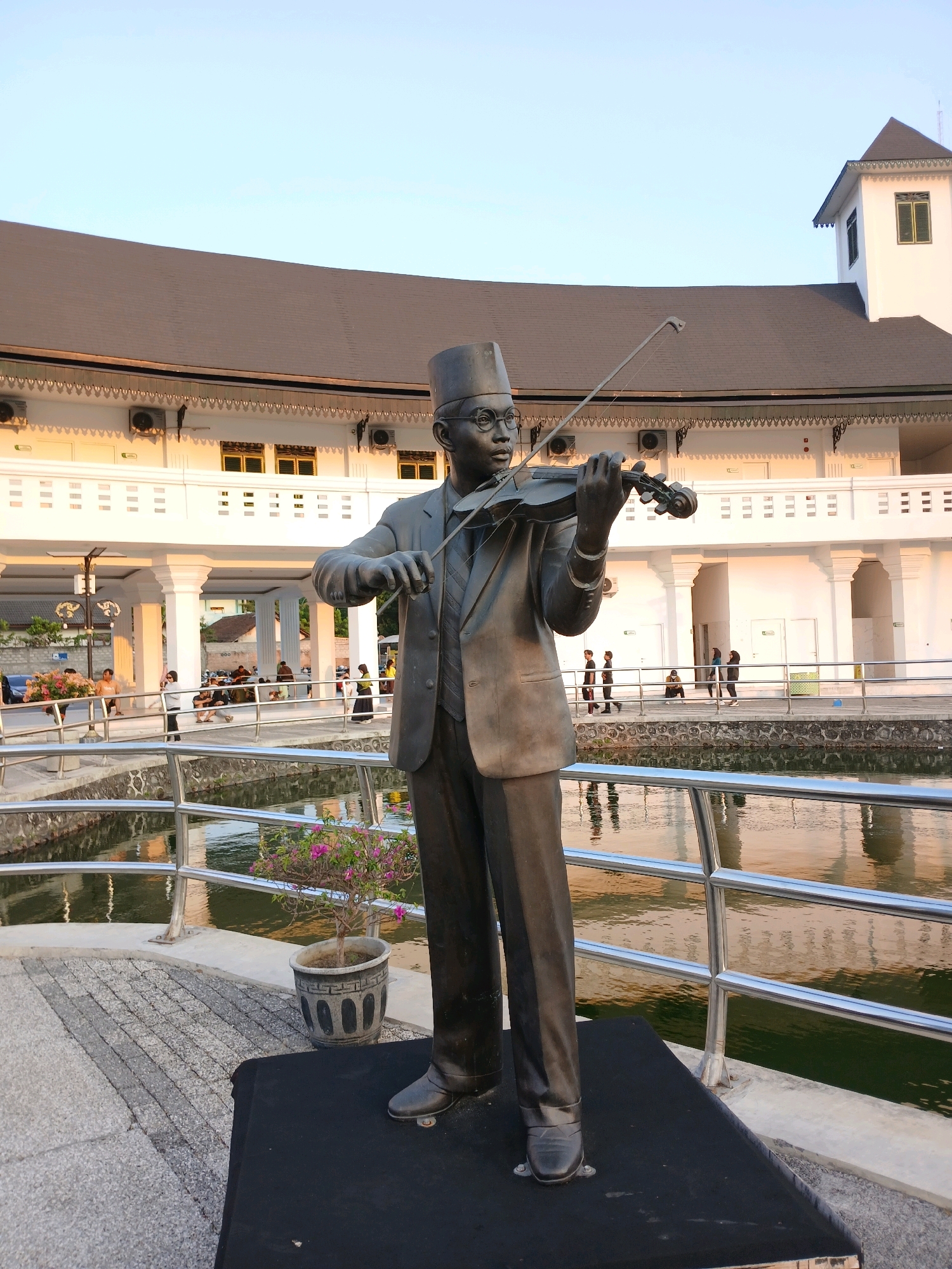 This is a full-shot, eye-level photograph of a bronze statue of a man playing the violin. The statue is positioned in the foreground, facing right, with the violin held up to his chin. He is wearing a formal suit, a tie, and a cap. Behind the statue, there is a body of water with a railing along its edge. Beyond the water and railing, a white, two-story building with a curved, dark roof can be seen. The building has several windows and air conditioning units visible on its exterior. In the distance, several people are walking and sitting near the building. The sky is clear and blue, suggesting it is daytime. The location is identified as the Taman Sari Water Castle in Yogyakarta, Indonesia.