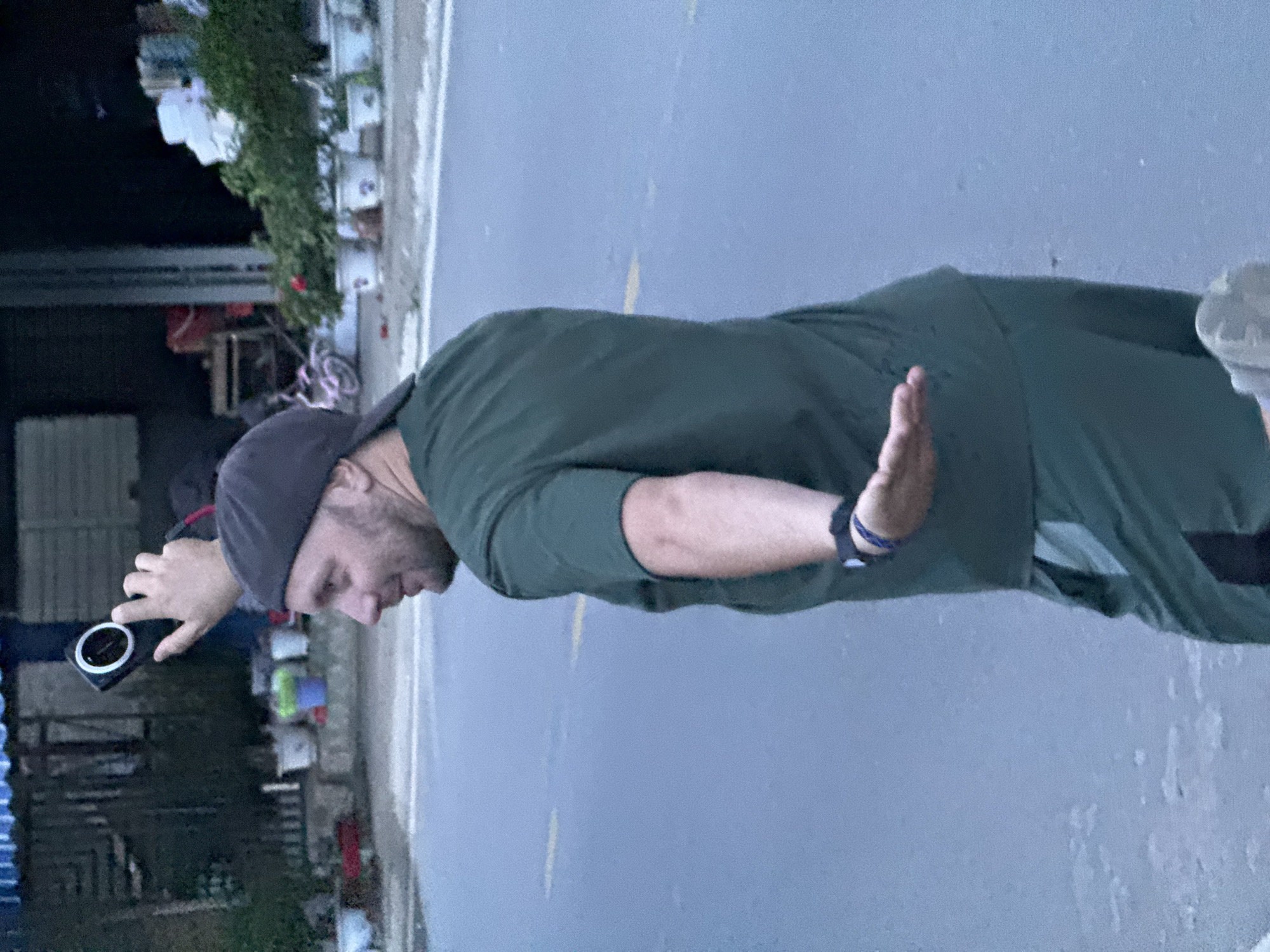 A man, appearing to be in his 30s or 40s, is captured standing outdoors, looking downwards and to his right. He is dressed in a dark green long-sleeved shirt and a dark baseball cap. In his left hand, he holds a black smartphone or a small camera, distinguished by a white ring around its lens, suggesting he is actively photographing or filming something at a low angle. His right hand is slightly raised with the palm facing forward, possibly in a gesture or for balance. A dark wristband is visible on his right wrist.
The man is positioned on a paved surface, likely an asphalt road or pathway, featuring faint yellow markings. To his left (relative to the viewer's orientation of the man), a strip of ground runs alongside with several white buckets and lush green foliage, possibly potted plants or dense bushes, indicating the presence of a building or wall. In the background, further simple structures and a bicycle are faintly visible. The lighting is soft and diffused, suggesting either an overcast day, early morning, or late afternoon/dusk. The overall setting implies a non-urban, possibly rural or semi-rural environment, fitting the context of Mù Cang Chải, Vietnam. There is no visible text in the image.