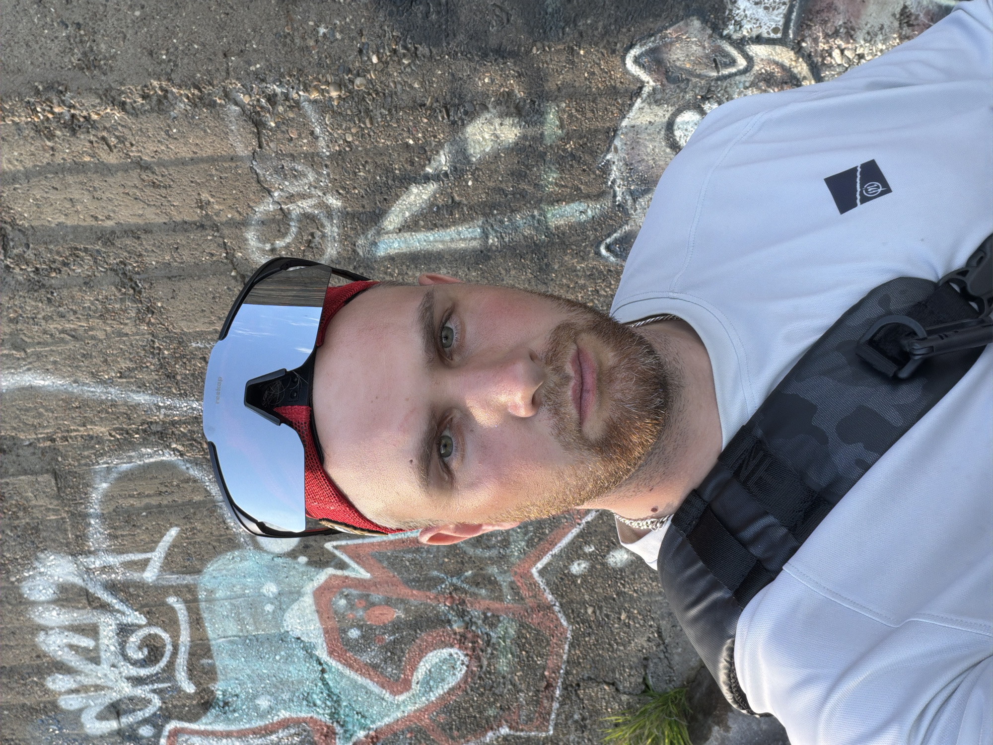 A medium shot of a man wearing sunglasses and a white long-sleeved shirt with a black patch on the upper left chest stands in front of a concrete wall covered in graffiti. He has short blond hair, a light beard, and appears to be wearing a black strap across his chest. The location appears to be in the city of Chaikovsky, Russia, as indicated by image origin.