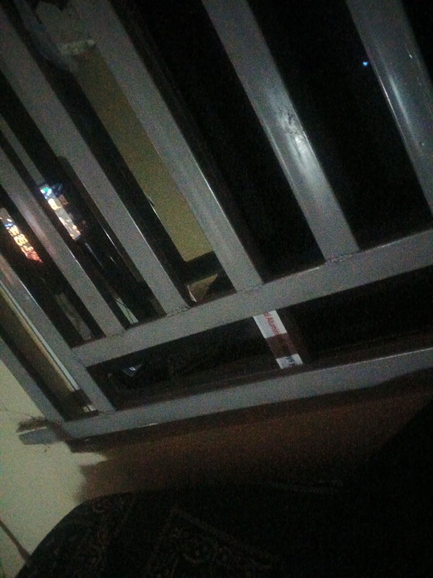 The image shows a close-up, angled view of a metal structure, possibly part of a ceiling or a shelving unit, with horizontal and diagonal metal bars. The lighting is dim, suggesting it might be nighttime or an indoor, poorly lit environment. In the background, through the gaps in the metal structure, faint reflections of lights are visible, hinting at an urban setting. One of the metal bars has a small white label with some red text, but it is illegible. The lower part of the image shows a dark, textured surface, possibly fabric. The scene is devoid of any identifiable people or clear actions. The location in Jalingo, Nigeria, cannot be determined from the visual information provided.