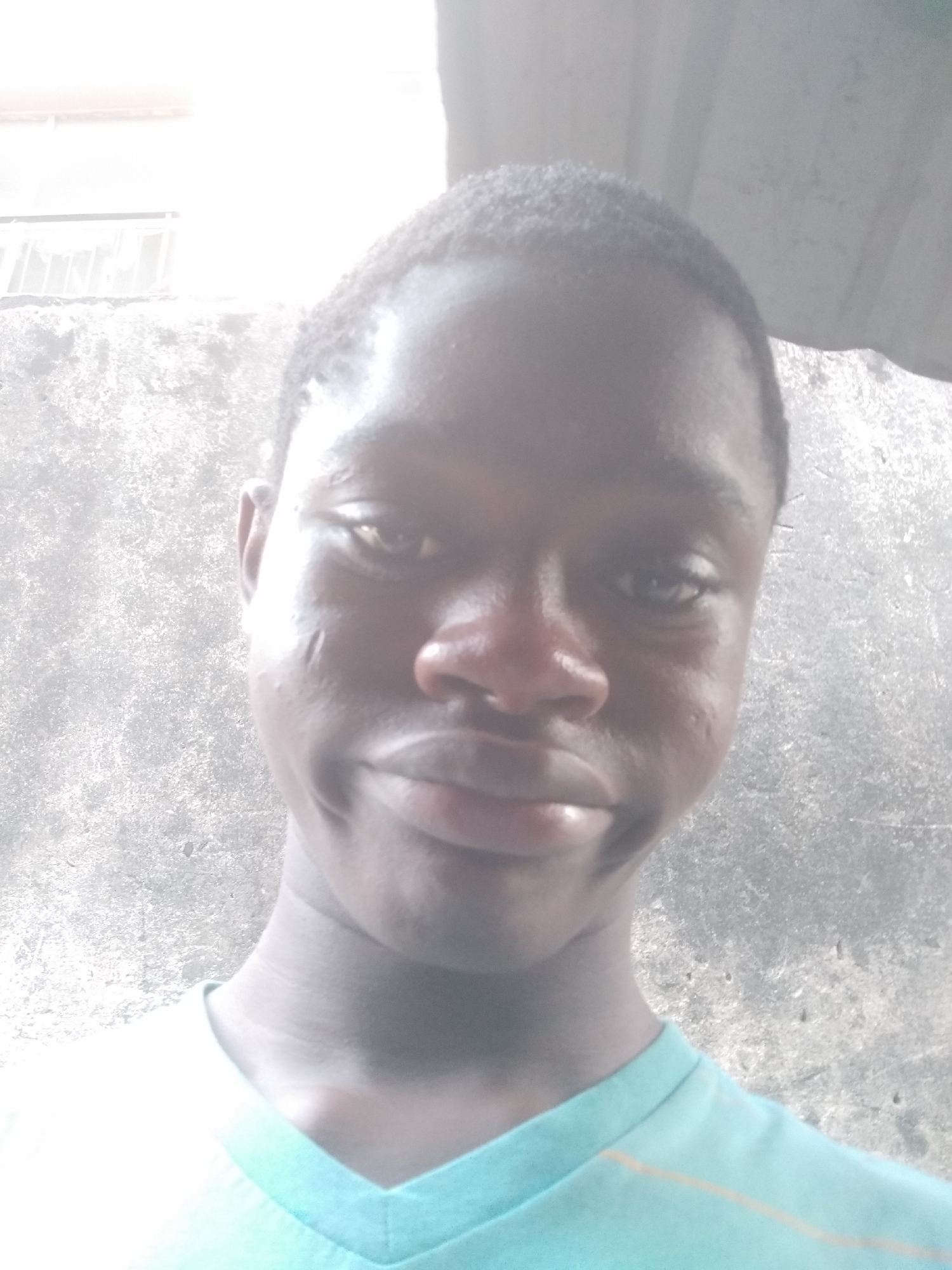 A young, dark-skinned person with short hair is centered in the frame. They are wearing a light blue v-neck shirt. The background shows a weathered concrete wall. The photo was taken in Shangisha, Nigeria.