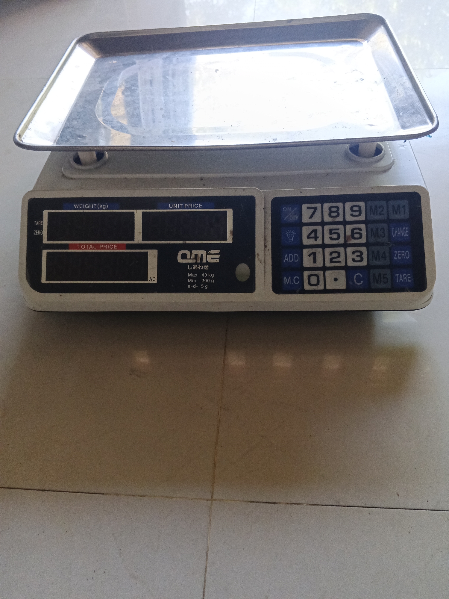 A digital scale rests on a tiled floor. The scale is made of white plastic and has a silver weighing tray. The scale has a digital display that shows "WEIGHT (kg)", "UNIT PRICE", and "TOTAL PRICE". There are also numerical buttons and function buttons labeled "ON/OFF", "ADD", "M.C", "M1", "M2", "M3", "M4", "M5", "CHANGE", "ZERO", and "TARE". The scale is positioned in what appears to be a market or shop setting, as indicated by the tiled floor, common in commercial establishments in Penambangan, Indonesia. No people are present in the image, and the scale is not in use. The environment is indoors, with overhead lighting reflected on the scale's tray.