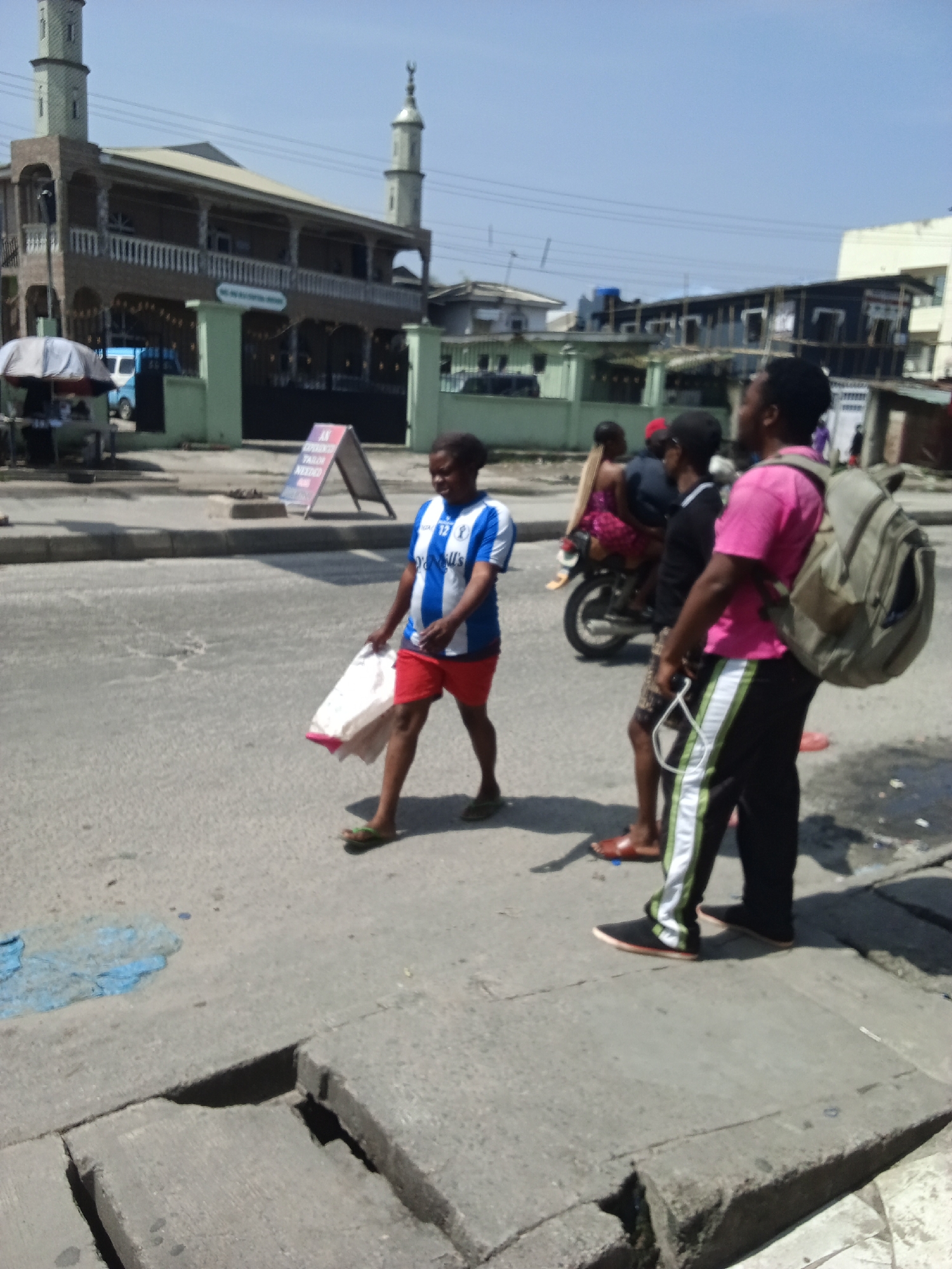 A group of people are standing and walking on a street in front of a building. A woman in a blue and white striped shirt and red shorts is walking and carrying a white bag. To her right, a man in a pink shirt and black pants with white stripes is standing. Behind them, a person is riding on a motorcycle. A building with two towers is visible in the background, and it appears to be a mosque. The street is paved with asphalt and there are some manhole covers and debris on the ground. The sky is clear and blue. The scene is likely in the city of Ado, Nigeria.