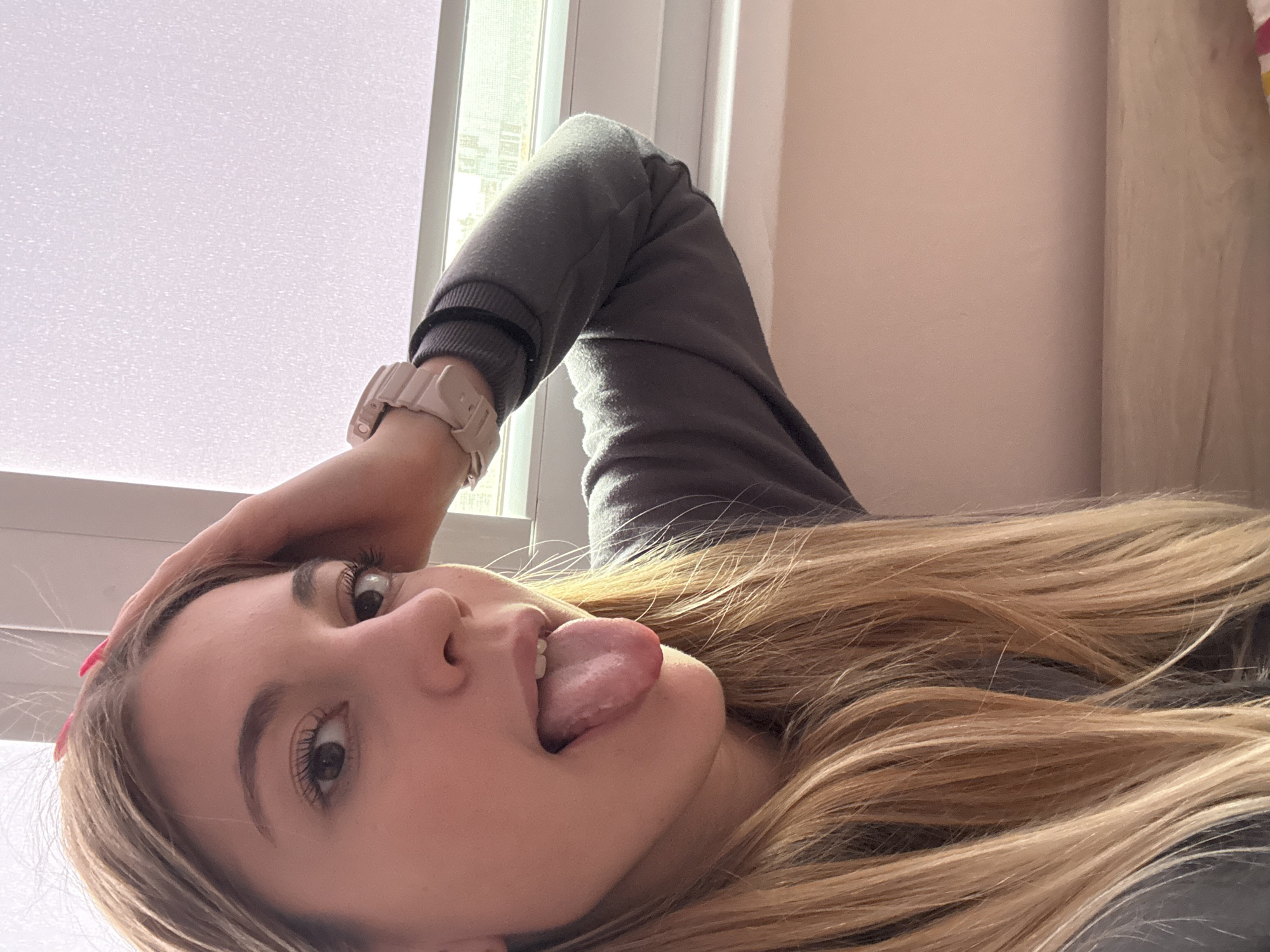 A young woman is captured in a close-up, low-angle indoor shot, playfully sticking out her tongue while looking directly at the camera. Her head is tilted, and her left hand rests casually on her head, revealing a hint of pink nail polish on one finger. She has long, straight, light brown to blonde ombre hair that frames her face and flows downwards. Her brown eyes are wide and bright, with visible mascara on her lashes. She wears a grey long-sleeved sweatshirt, and a light pink or beige digital watch is prominent on her left wrist.
The background is softly illuminated by a large, bright window on the left, possibly featuring frosted glass or blinds, which diffuses the daylight. To the right, a plain light pink or beige wall provides a warm backdrop. The bright, even lighting suggests it is daytime. The woman's expression is playful and mischievous. There is no visible text in the image. The provided location context for this media is Sofia, Bulgaria.