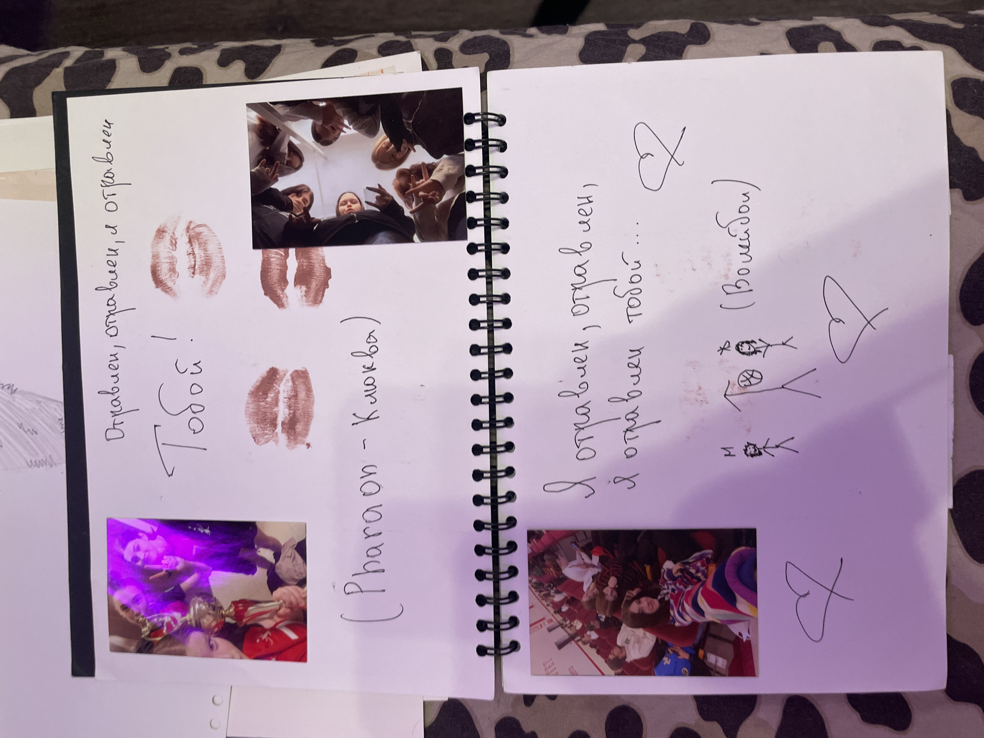The image shows a scrapbook or journal with two pages visible, laid out on a patterned surface.
The top page features a photograph on the left of a group of young people, one of whom is holding a trophy and making a peace sign. To the right of the photograph, there is handwritten text in Russian: "Отравиен, отравлен, я отравлен" which translates to "Poisoned, poisoned, I am poisoned". Below this, more Russian text reads "Тобой!" which translates to "By you!". There are also two lipstick kiss impressions on this page. At the bottom of the top page, a title is written in English and Russian: "(Pharaon - Клюква)".
The bottom page features another photograph on the left showing a young woman wrapped in a colorful blanket, looking towards the camera. To the right of this photograph, more Russian text is handwritten: "Я отравлен, отравлен, я отравлен тобой..." which translates to "I am poisoned, poisoned, I am poisoned by you...". Below this, there are two simple stick-figure drawings: one of a person and a basketball hoop, and another of a person with an asterisk above their head. Next to these drawings, in parentheses, is the Russian word "(Волейбол)", which translates to "Volleyball". Two heart shapes are also drawn on this page.
The image does not contain any information that would allow for the determination of its location in the city of Kokkorevo, Russia.
