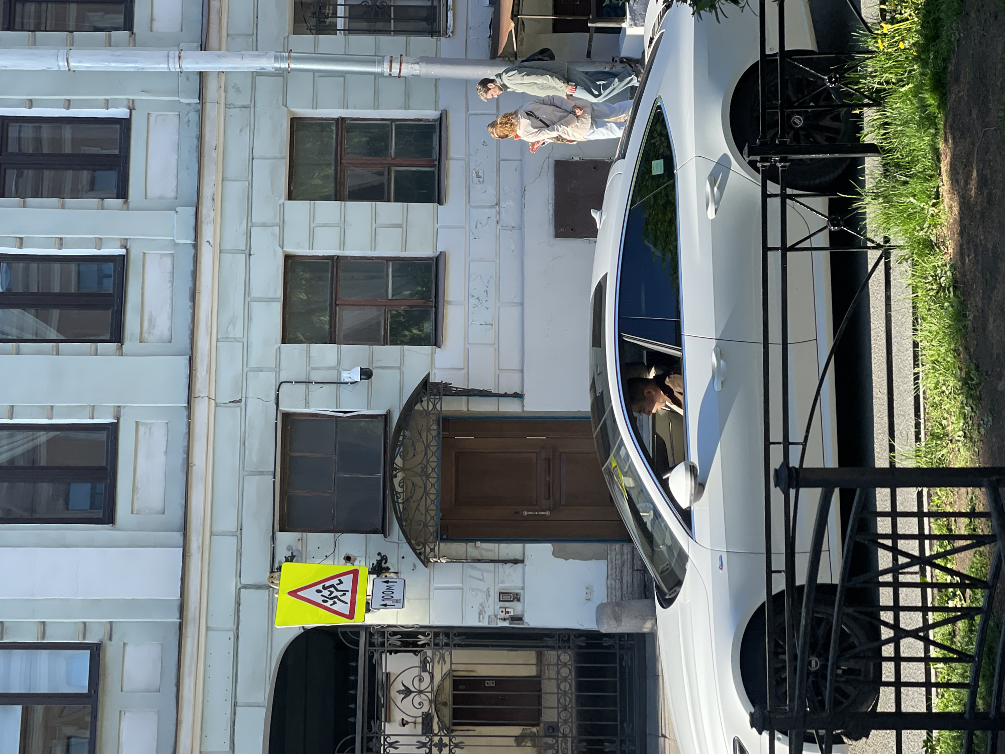 The image shows a building facade in Saint Petersburg, Russia, next to a parked white car. The building has a light blue exterior with a series of windows with dark frames and a central wooden door covered by a wrought-iron awning. A pedestrian crossing sign and a distance sign are affixed near the gate in front of the entrance. Two individuals are standing in front of the building, likely observing something above. A black metal fence runs parallel to the car, and a grassy area is located to its side. The image is slightly rotated clockwise.