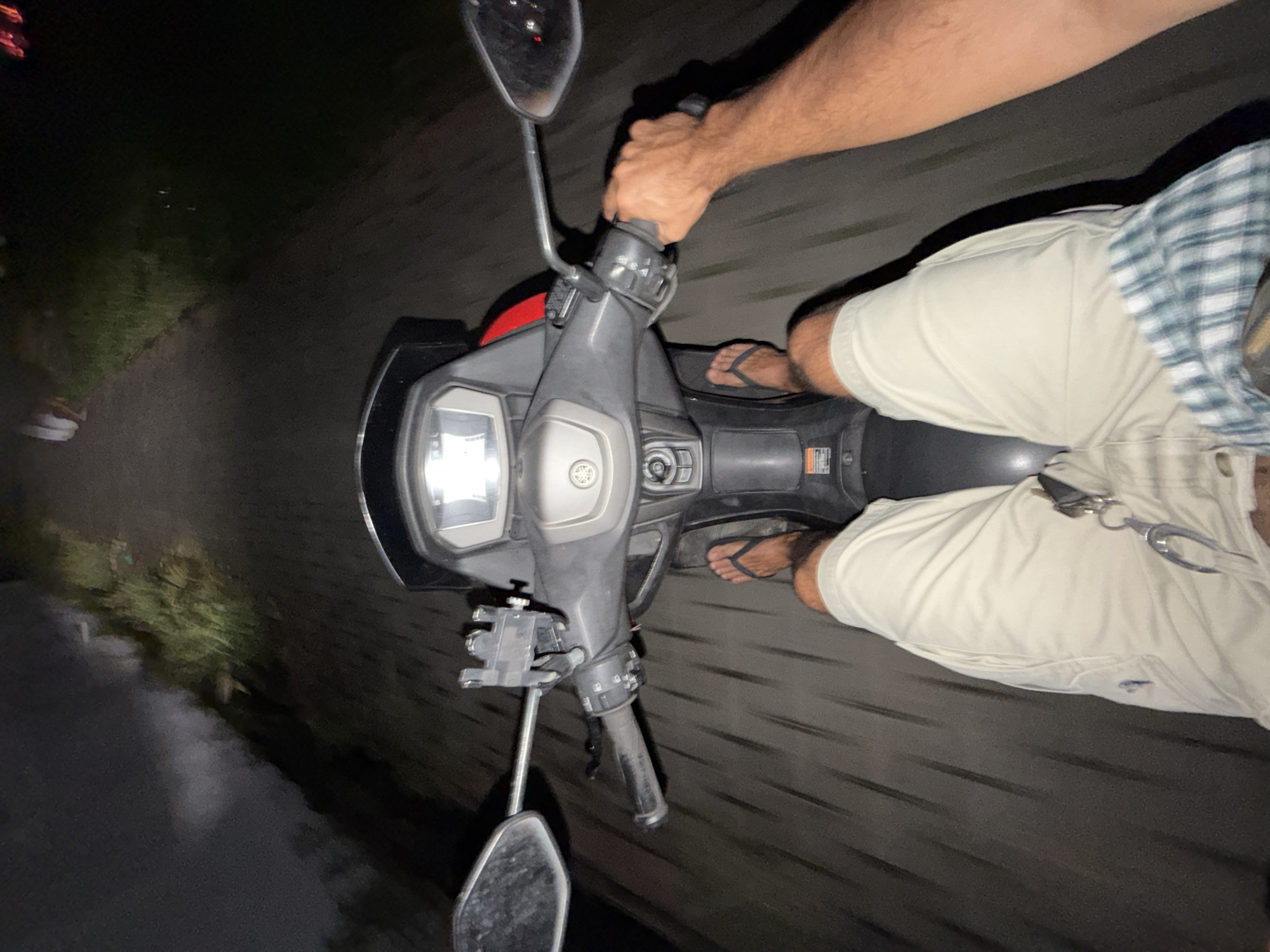 Captured from the rider's perspective, the image depicts a person operating a scooter on a paved road. The rider is seen wearing light-colored shorts, a plaid shirt, and flip-flops. Their left hand is gripping the handlebar, while their feet rest on the footrests. The scooter features a front headlight and mirrors. The surrounding environment suggests a dark, possibly nighttime, setting with roadside vegetation. The location is North Kuta, Indonesia.