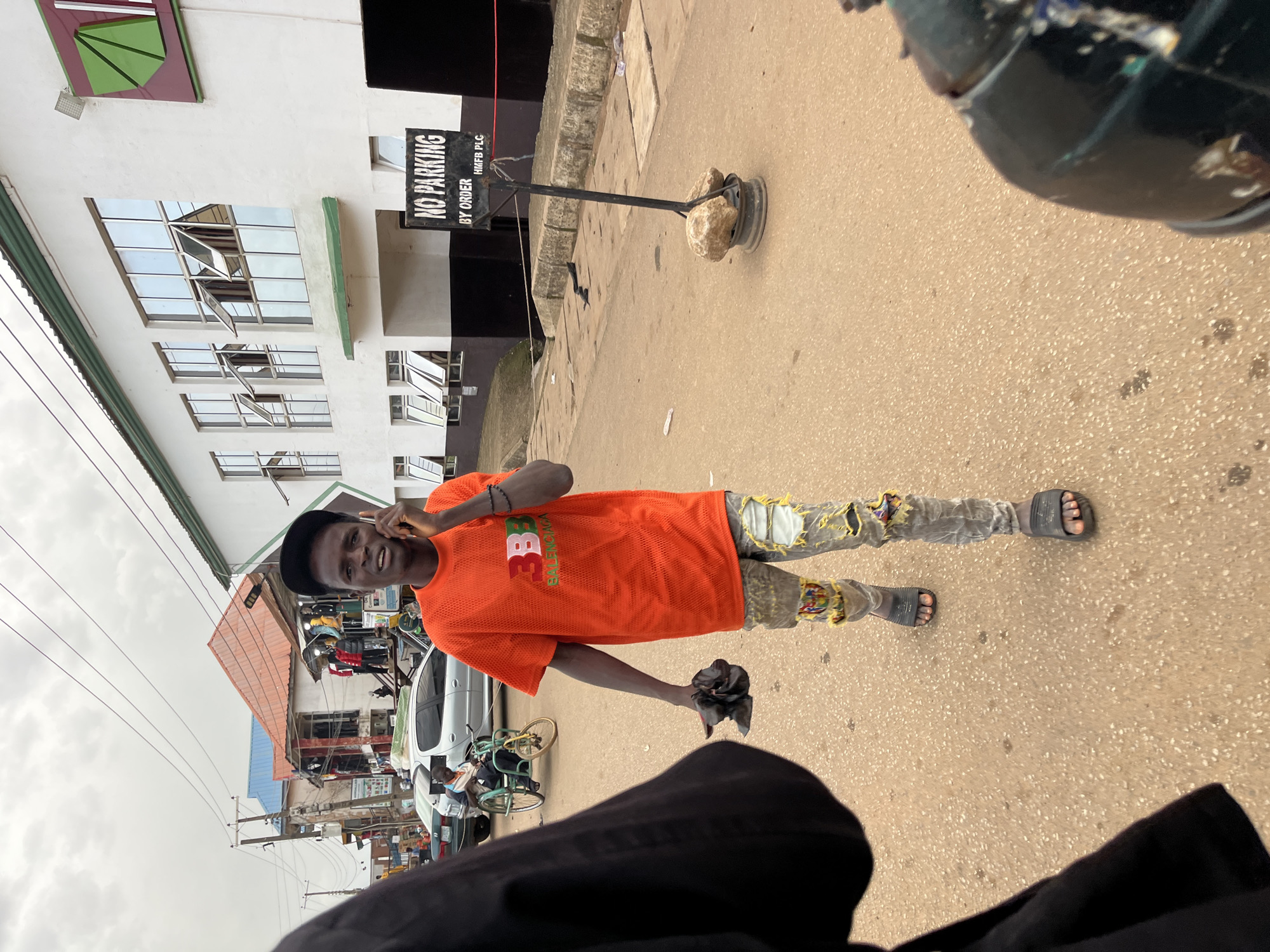 In Bukuru, Nigeria, a young Black male, wearing a bright orange mesh t-shirt with "3B3" and "BALENCIAGA" printed on it, ripped grey jeans, and black slide sandals, is captured mid-stride on a paved street. He is holding a black mobile phone to his right ear, appearing to be engaged in a conversation. In his left hand, he clutches a crumpled black object. Behind him, several cars are parked and moving, including a silver sedan and a green tricycle with a passenger. In the background, a two-story building with white walls and numerous reflective windows stands prominently. A "NO PARKING" sign, supported by a metal pole and secured with rocks, is positioned on the side of the road. The sky is overcast, suggesting a cloudy day. The overall scene depicts a typical street view in a Nigerian urban environment, with elements of daily life, commerce, and transportation.