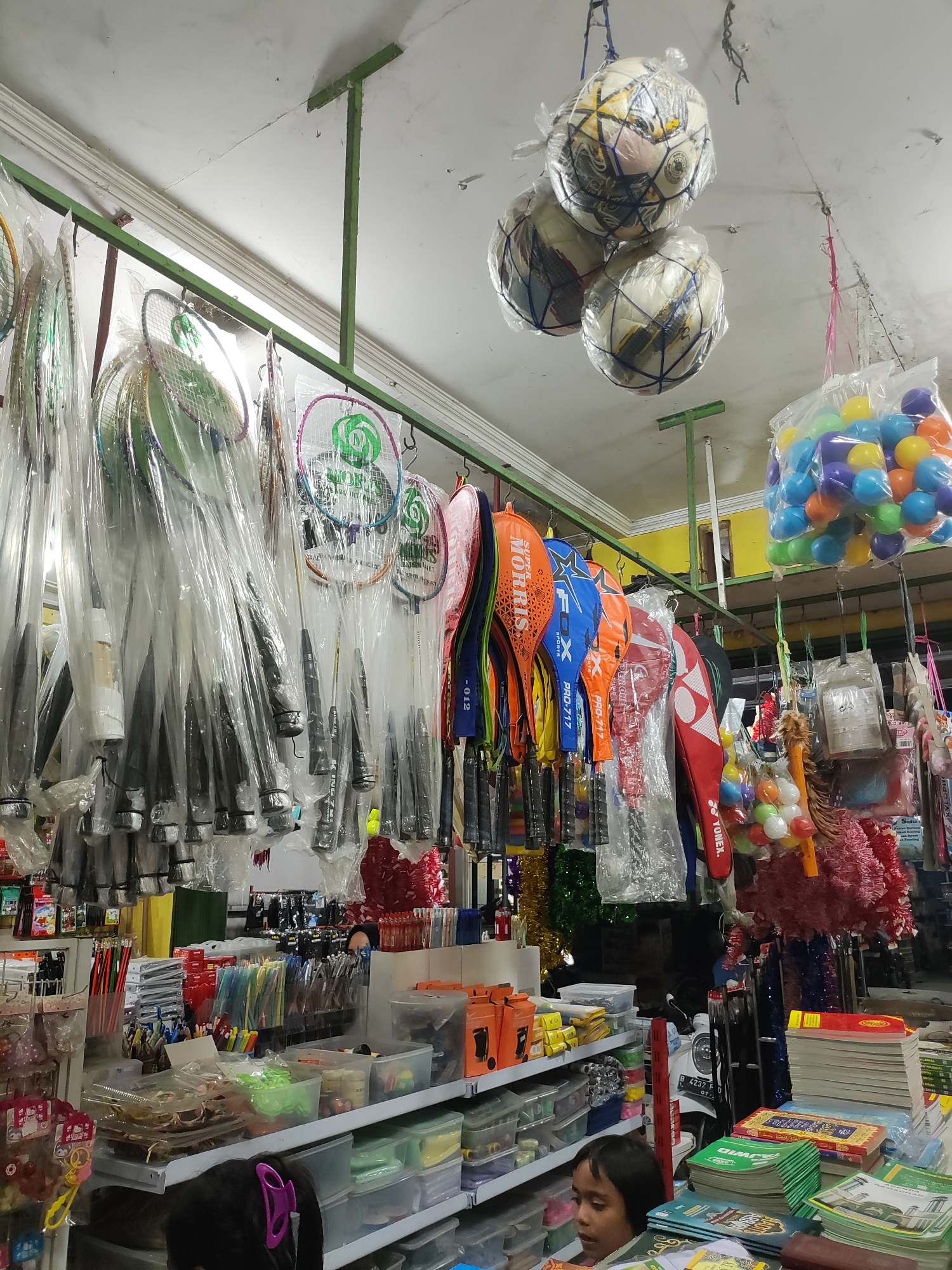 The image depicts the interior of a small shop in Wanajaya, Indonesia, likely a convenience store or a general goods store. The ceiling is white with exposed green metal supports. Hanging from the ceiling are three white and colored soccer balls, encased in plastic netting. To the right, a clear plastic bag is filled with colorful small balls, predominantly blue, green, and yellow.
Dominating the mid-ground are numerous badminton rackets, individually wrapped in clear plastic. They are hung in neat rows from a green metal rack. Some rackets are brightly colored with prominent logos such as "MORRIS" and "FOX". Below the rackets, shelves are stocked with various items. These include boxes of stationery, such as pens and pencils, neatly arranged. Other shelves hold clear plastic containers filled with more small, colorful balls and other small items.
In the foreground, a young child with dark hair and wearing a pink hair clip is visible from the chest up, looking to the right. The child is positioned in front of a shelf stacked with books. The books are predominantly green and red and appear to be religious texts, with Arabic script visible on their covers. Other items like stacks of paper and various containers are also present on the shelves. The overall impression is that of a busy, well-stocked retail environment.