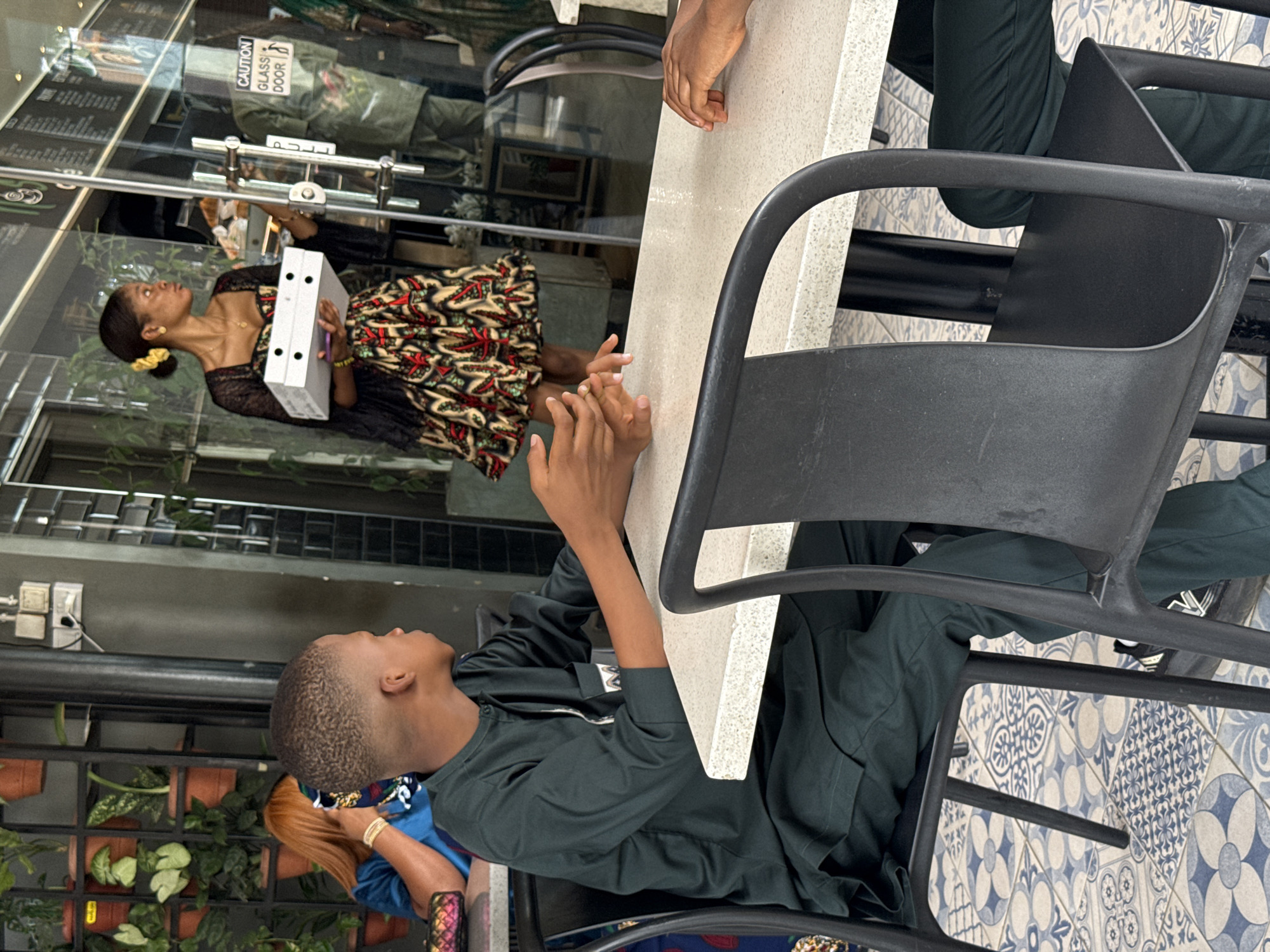 Here's a forensic description of the image:
**Location:** The image appears to be taken inside or near a commercial establishment, likely a restaurant or cafe, in Abuja, Nigeria. This is based on the presence of potted plants visible near the entrance, the tiled flooring resembling local designs, and the architectural style.
**People:**
* Two individuals are seated at a table. One appears to be a young male, with a short haircut, wearing a dark green garment. The other person is only partially visible, showing only their arm.
* Reflected in the glass entrance is a woman carrying a white rectangular box. She is wearing a patterned dress with black lace details.
**Objects:**
* A rectangular table with a light-colored surface.
* Two black chairs are visible at the table.
* A white box held by the woman in the reflection.
* Potted plants near the entrance.
* A glass door with a "Caution Glass Door" sign.
* Tiled flooring.
**Actions and Interactions:**
* The two individuals at the table seem to be in conversation, or waiting.
* The woman is entering or exiting the establishment, carrying a box.
**Environment:**
* The setting is a commercial area, likely during daylight hours, as suggested by the light.
* The area seems to be relatively clean and well-maintained.
* The style and clothing suggest a location in Nigeria, specifically Abuja.