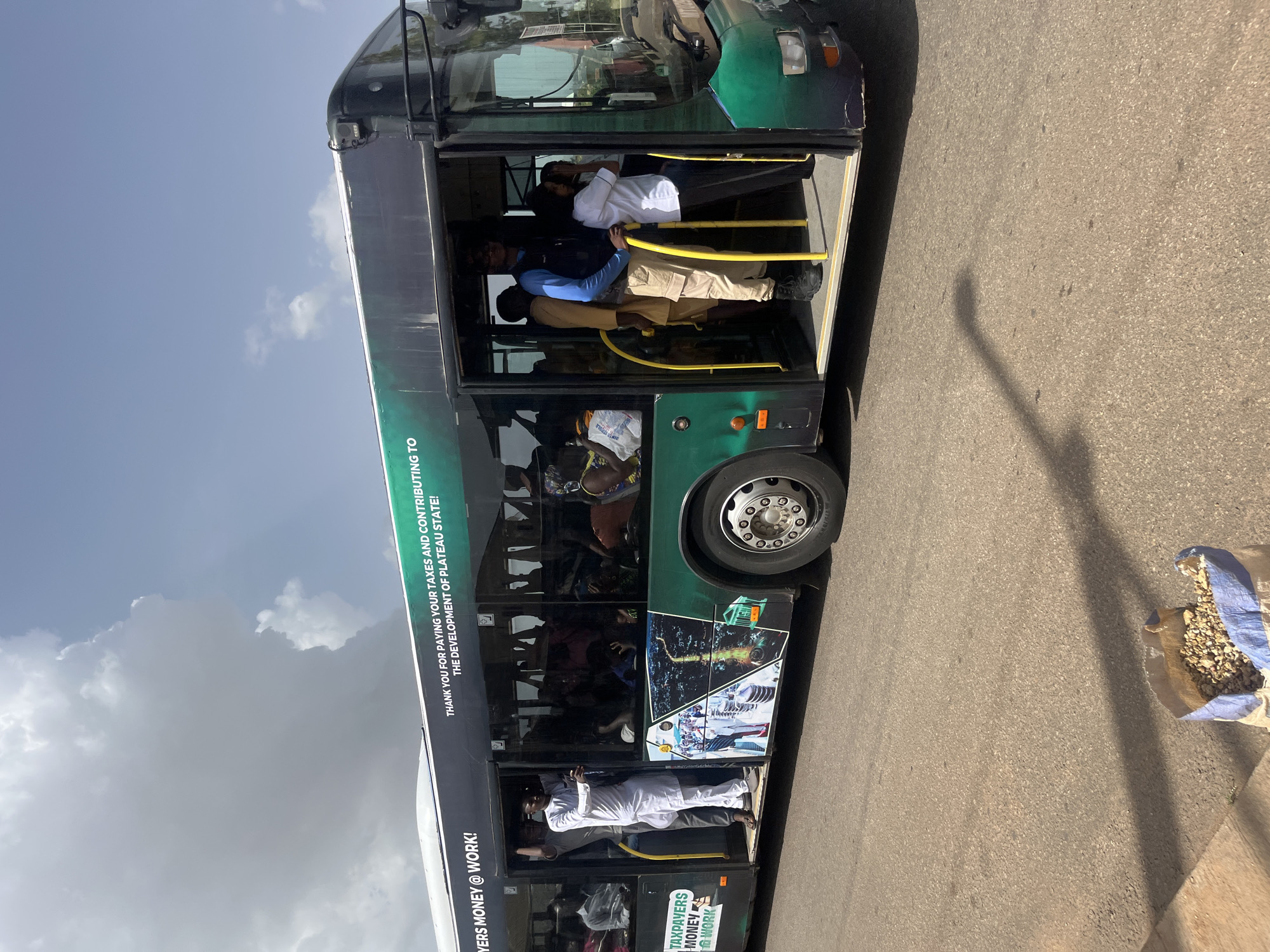 An image shows a full bus with many people inside. The bus is dark with a green stripe on the side that reads "THANK YOU FOR PAYING YOUR TAXES AND CONTRIBUTING TO THE DEVELOPMENT OF PLATEAU STATE!". On the front left side of the bus, there is text that says "TAXPAYERS MONEY @ WORK!". The bus appears to be in motion on a paved road. There is a bag of gravel on the ground in the foreground. The sky is cloudy. The image appears to be taken in Gura Top, Nigeria.
